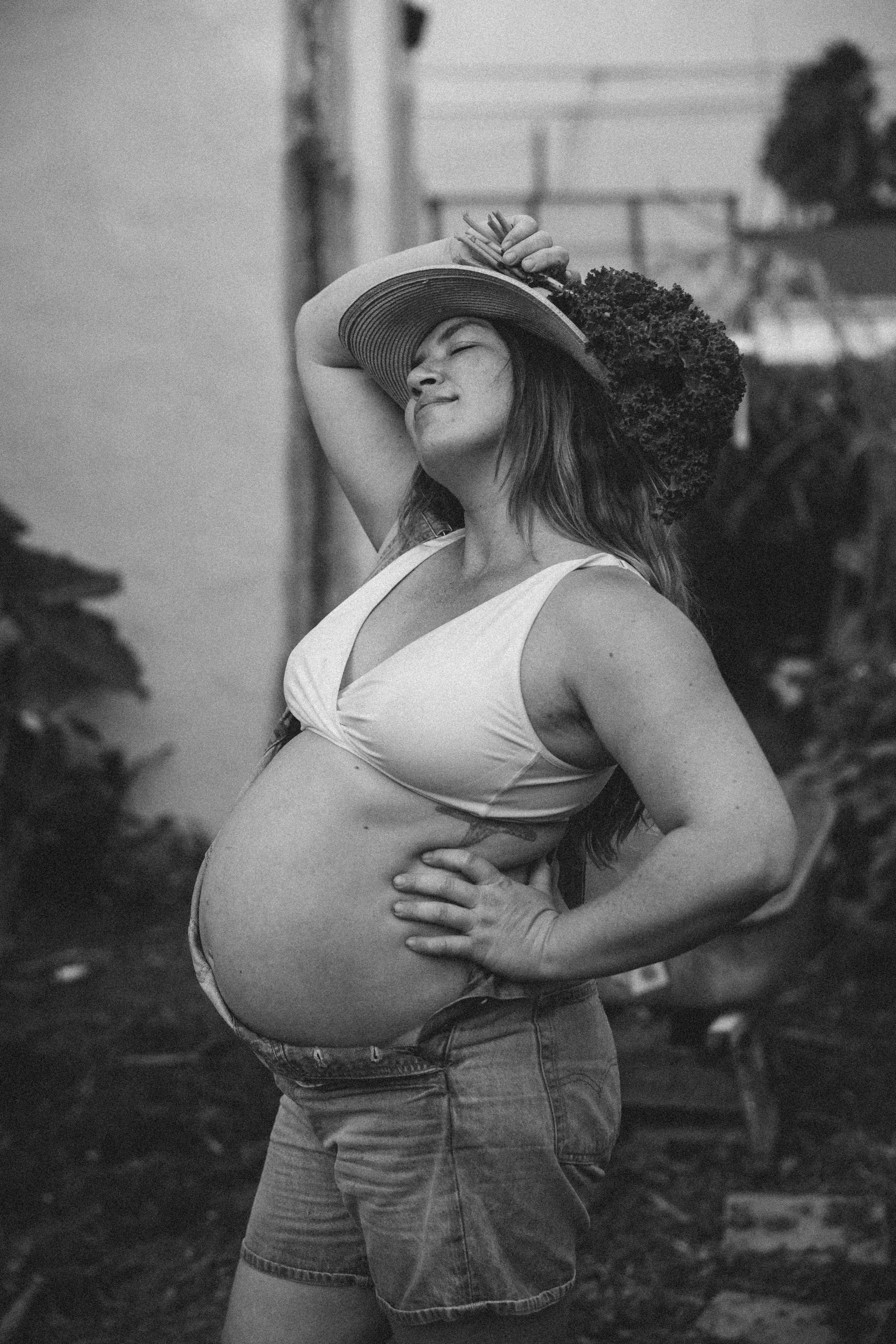 Pregnant woman wearing a wide-brimmed hat and a sleeveless top, standing outdoors with her hand on her hip, squinting towards the sunlight.