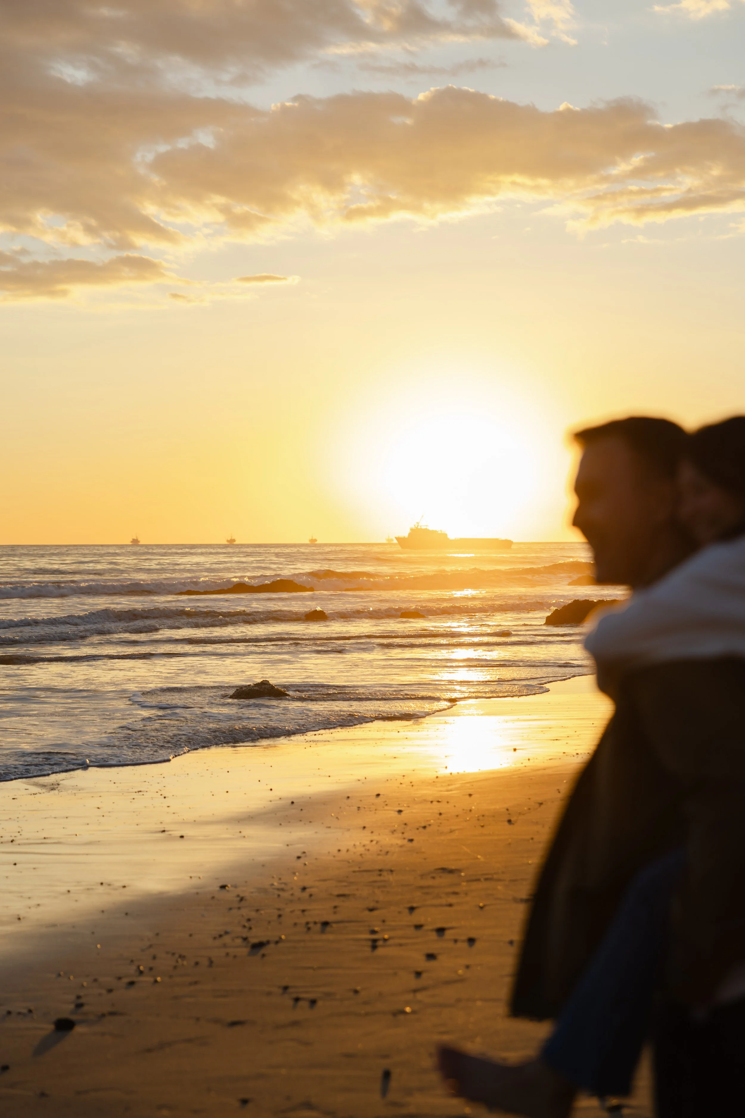 People walking along a sandy beach during sunset with the ocean and ships in the background.