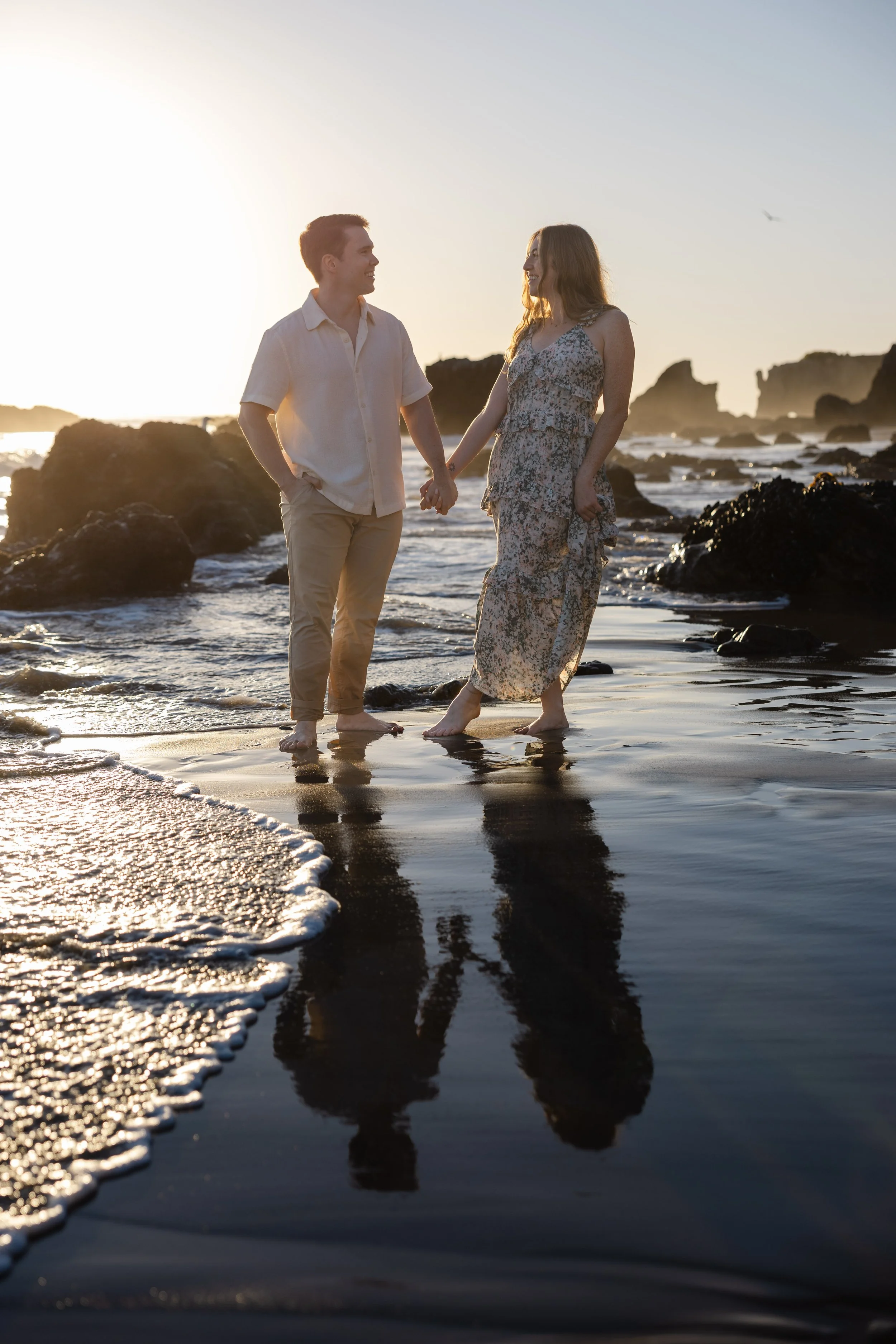 A couple holding hands and walking along the beach at sunset, barefoot, with rocky formations in the background.