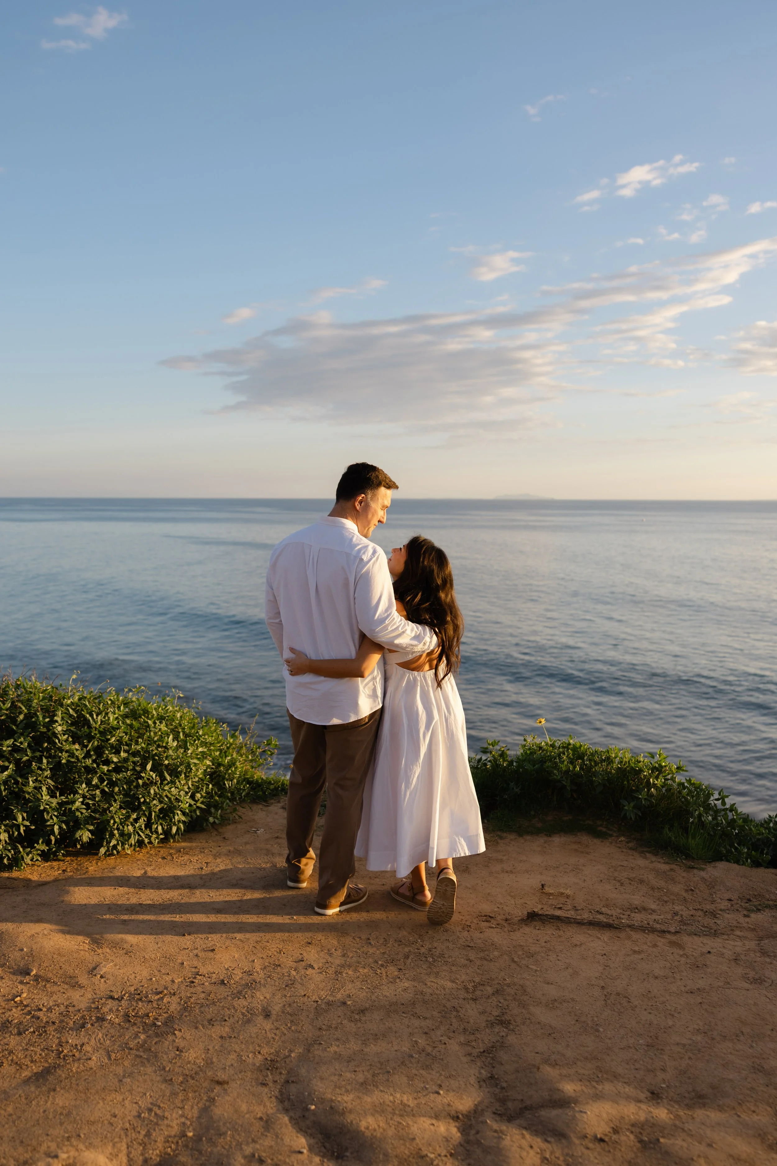 A couple walking together near the ocean during sunset, with the man dressed in a white shirt and the woman in a white dress.