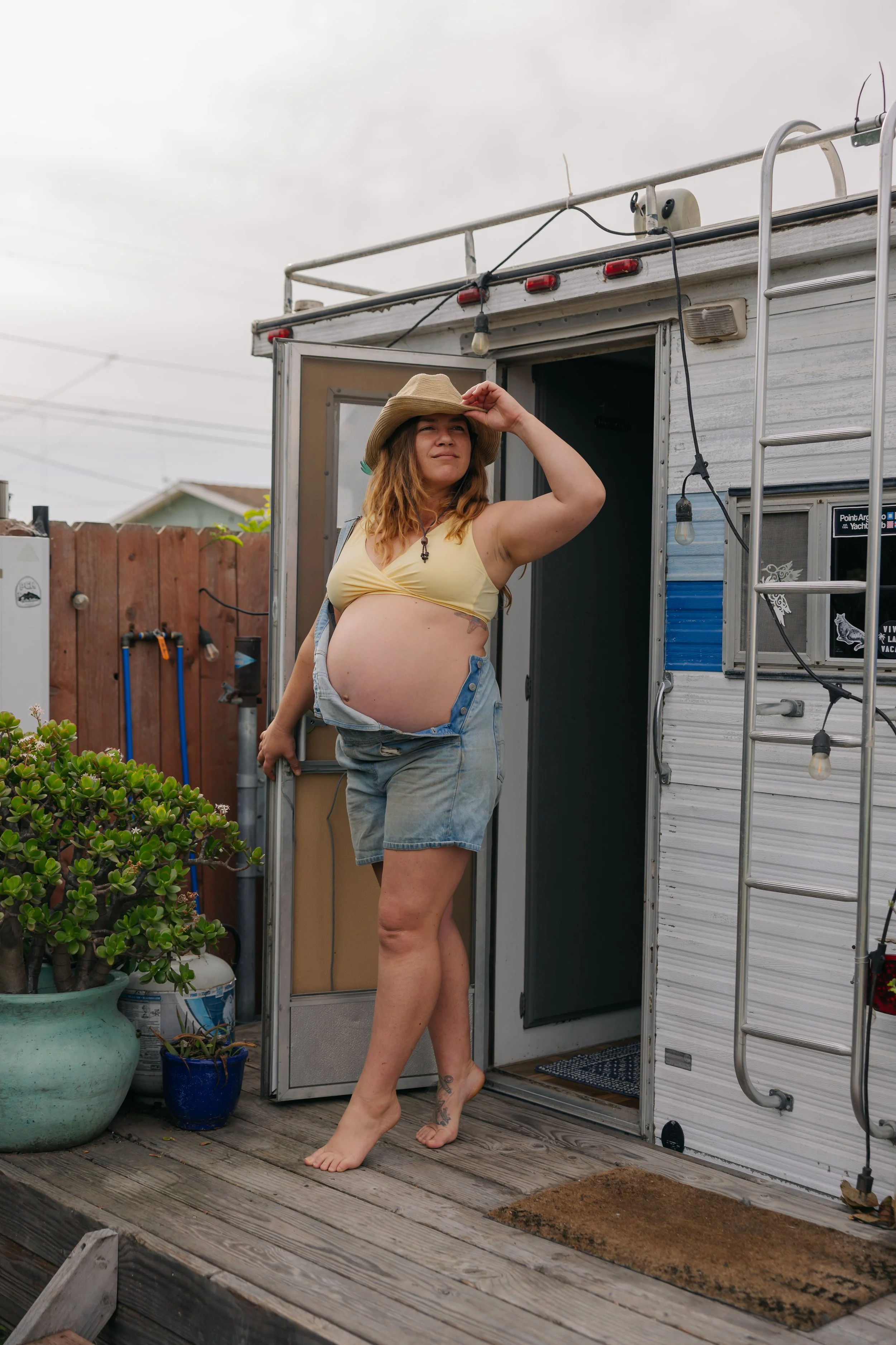 A pregnant woman standing barefoot on wooden deck, wearing denim shorts and a yellow top, holding a straw hat, standing at the entrance of a small camper or tiny house.