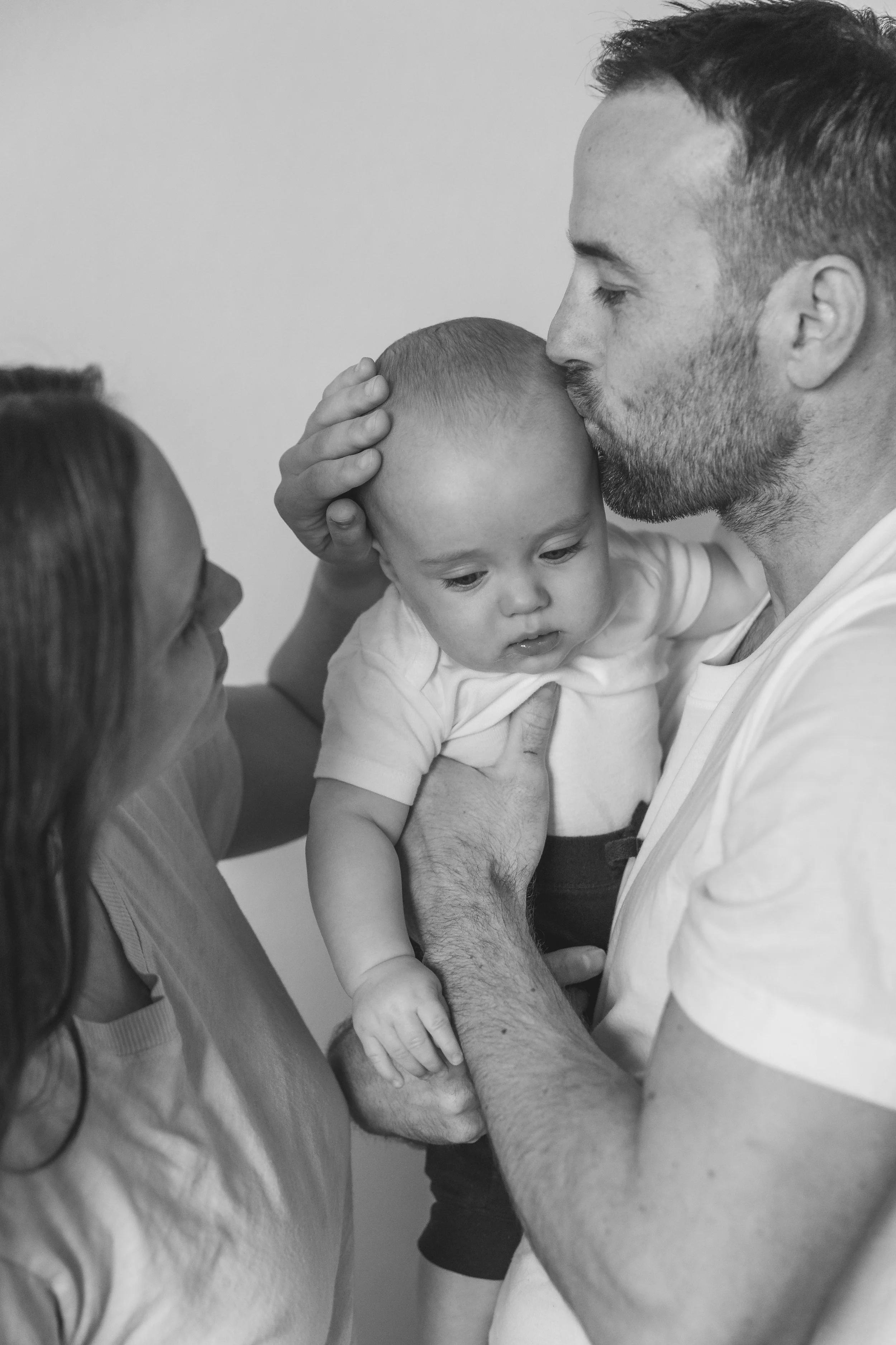A black and white photo of a family with a father and mother holding their young child. The father is kissing the child's forehead while the mother looks at them with a gentle expression. The father's hand supports the child's chest, and the mother h