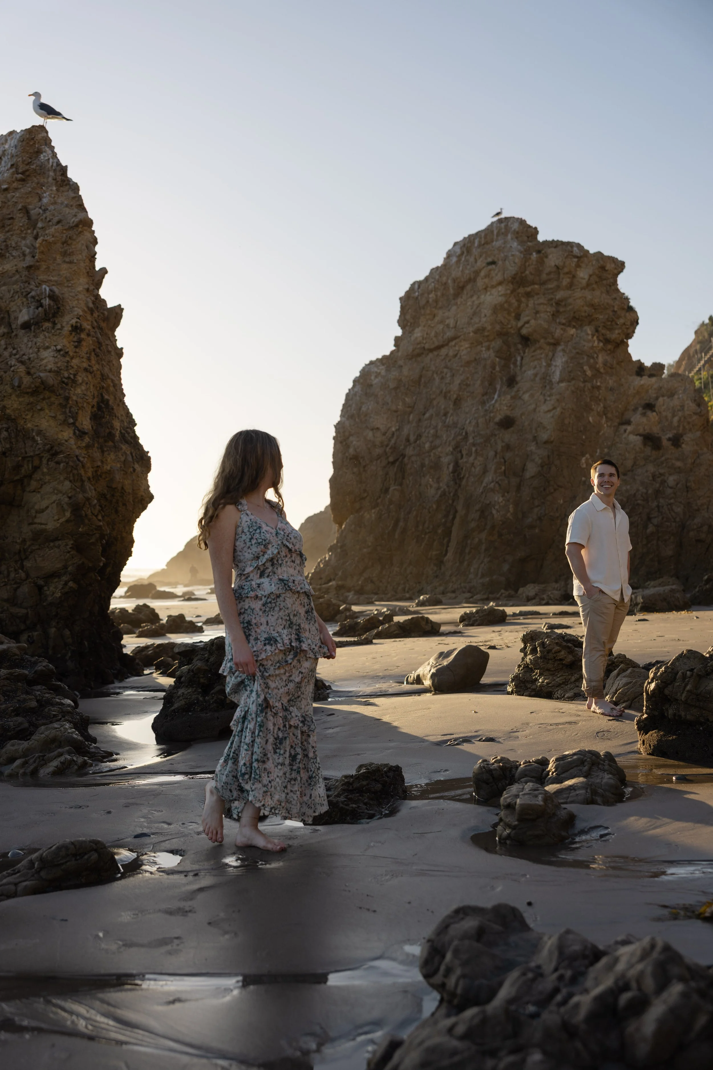 A couple walking barefoot on a sandy beach with large rocks and cliffs around them during sunset, one woman looking at the ocean and a man smiling.