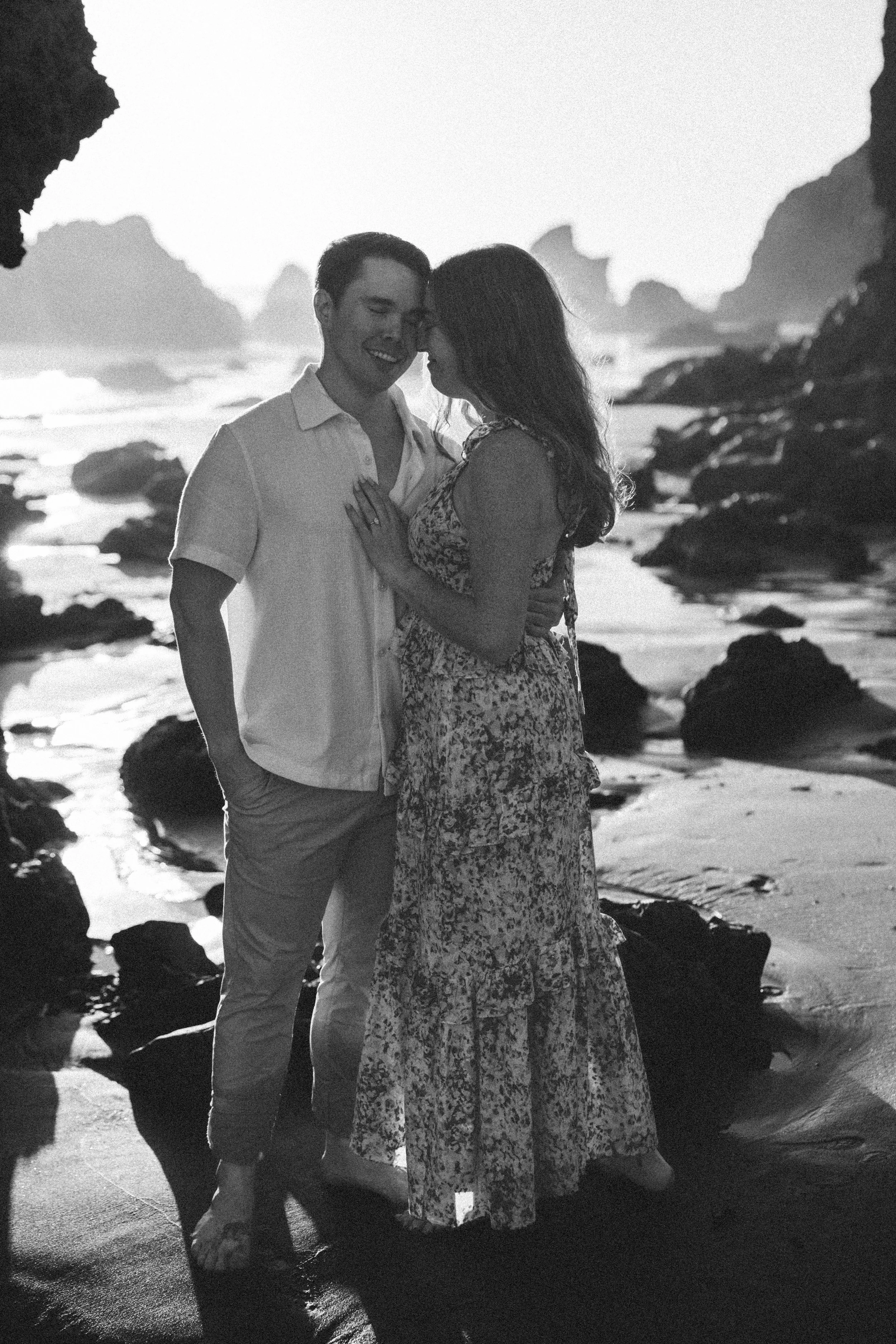 A black and white photo of a couple standing on a beach at sunset, close together, with the woman's hand on the man's chest, sharing an intimate moment.