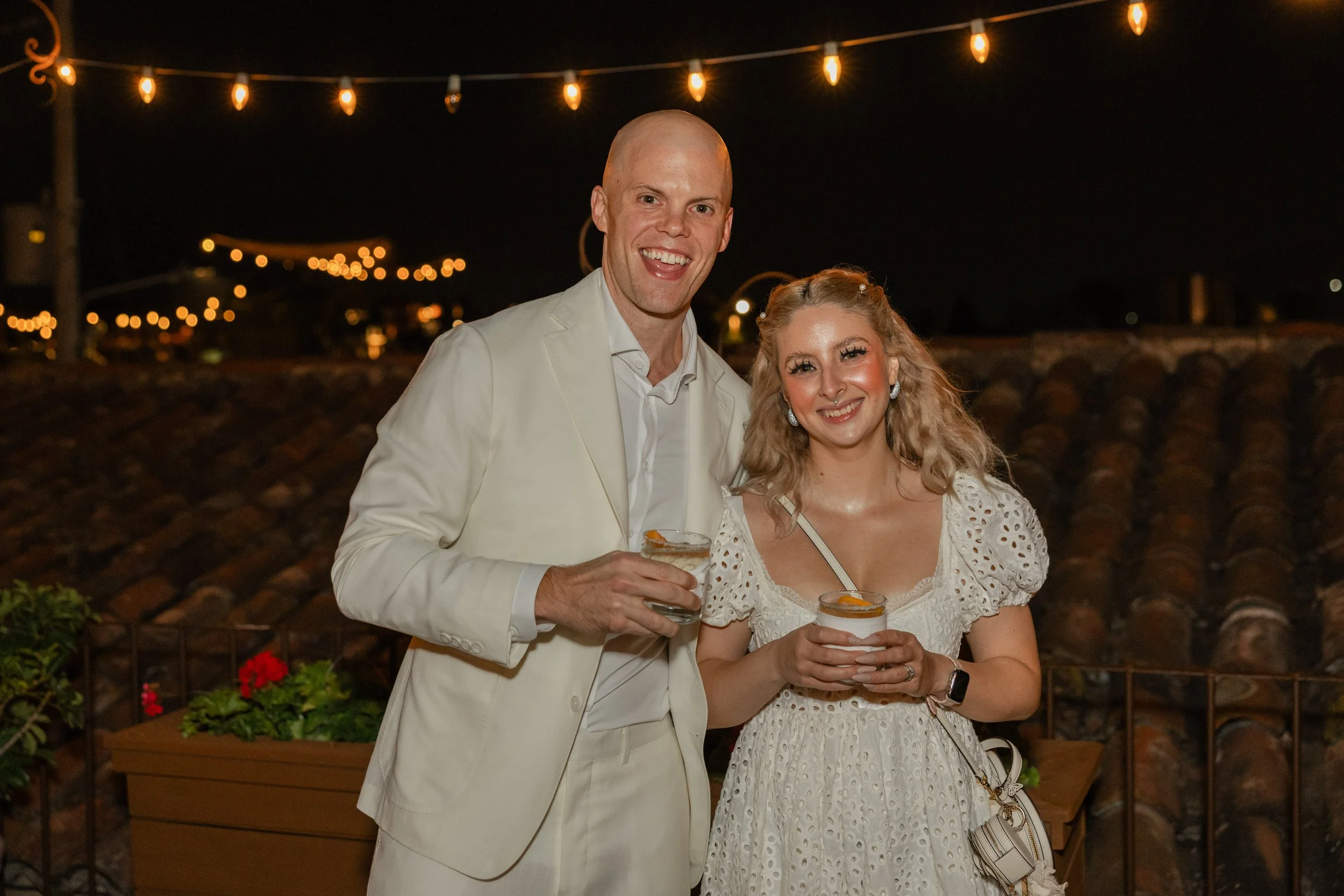 A man and woman smiling at night, holding drinks, standing outdoors with string lights above and a tiled roof in the background.