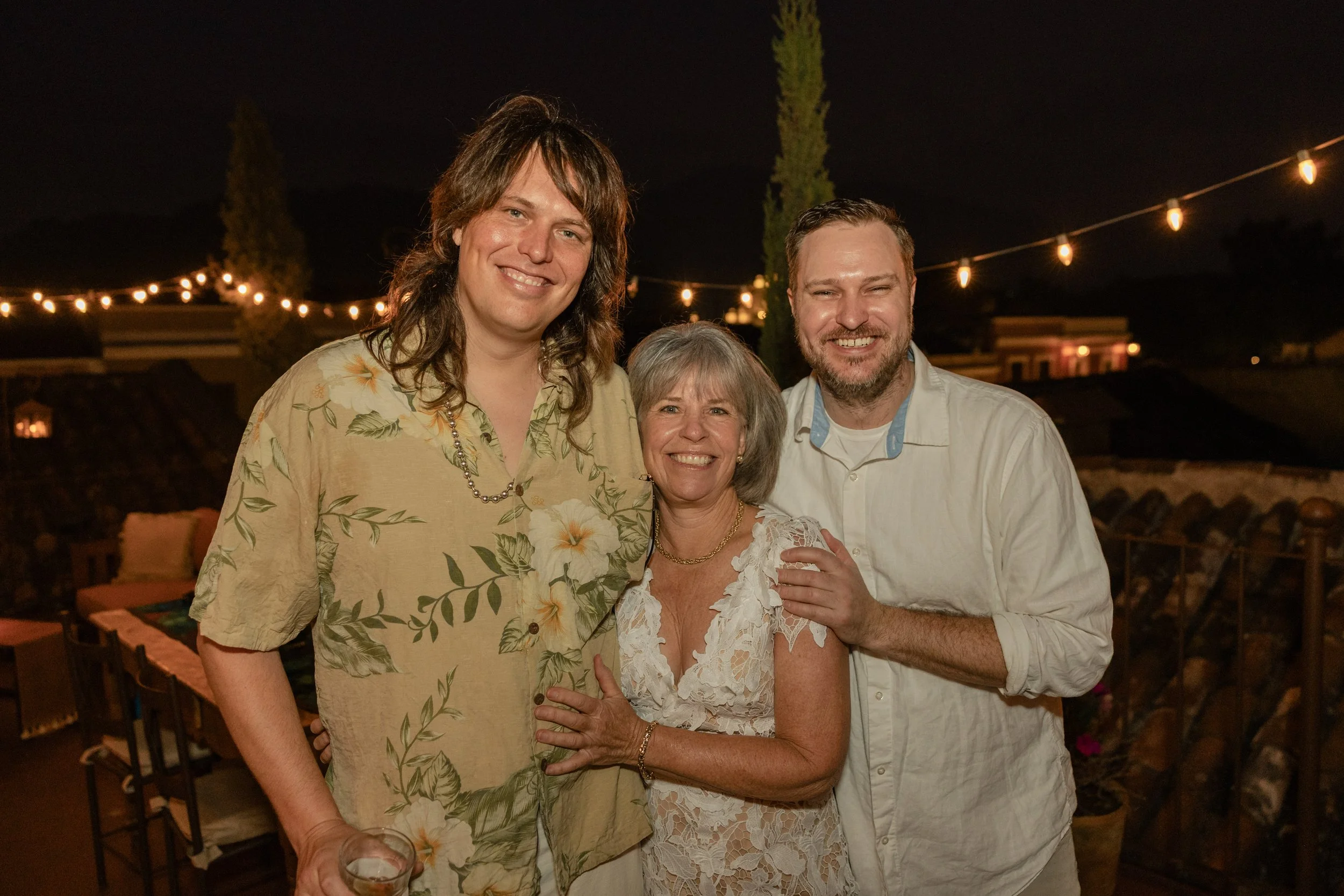 Three smiling people, two men and one woman, standing closely together at night outdoors, with string lights and rooftops in the background.