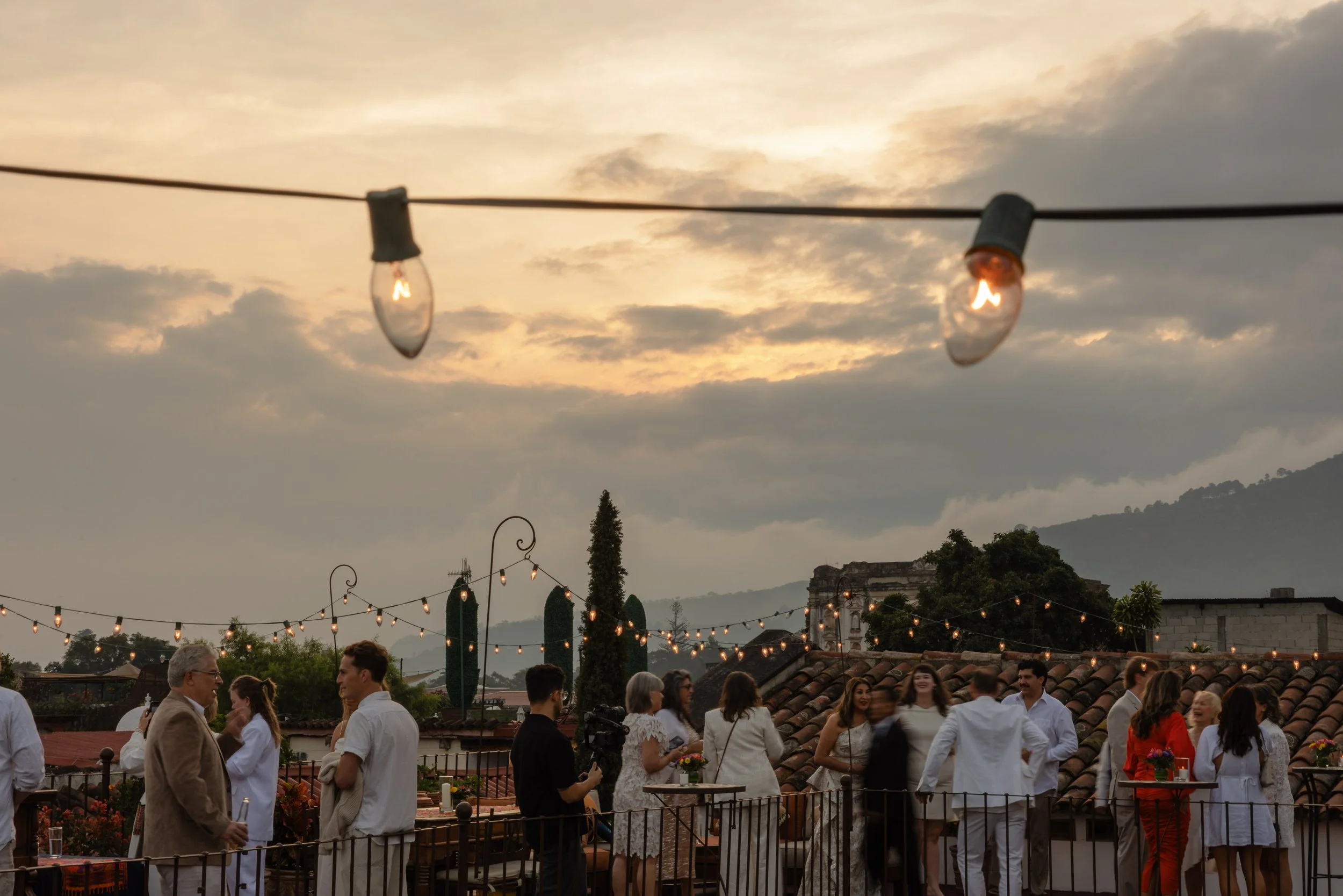 People socializing on a rooftop terrace during sunset, with string lights hanging overhead and mountains in the background.