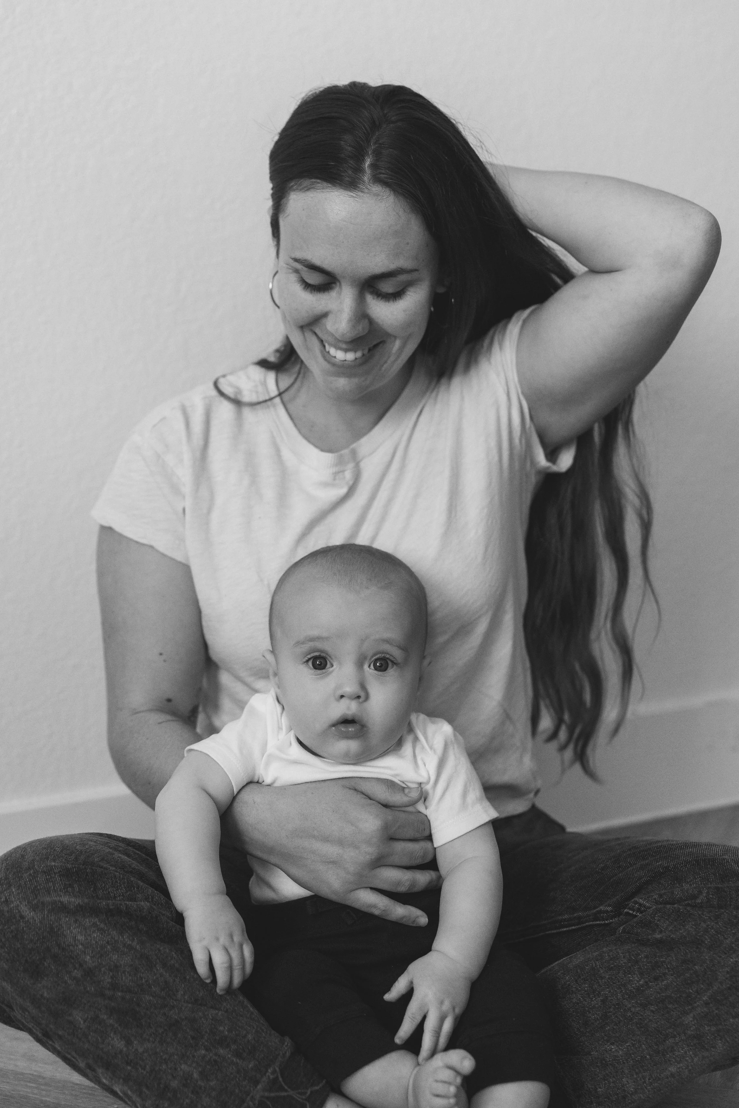 Black and white photo of a woman sitting on the floor with a baby on her lap, both looking at the camera, woman smiling with her hand behind her head.