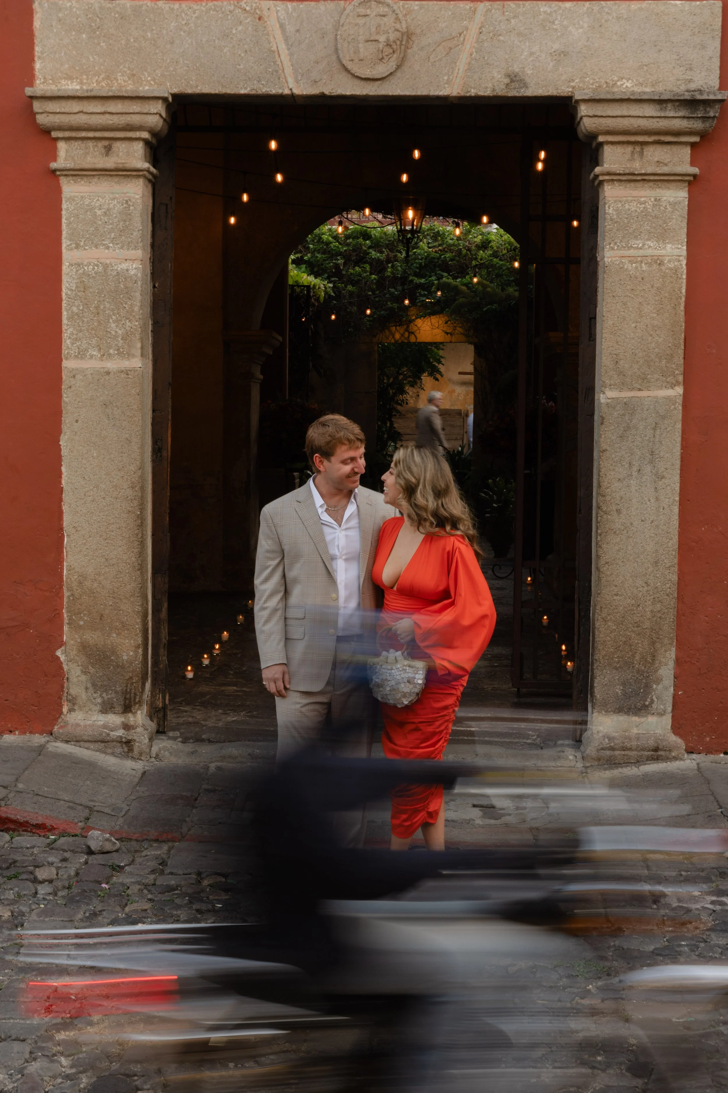 A man and woman look at each other and smile in front of a historic building entrance with hanging lights, while a blurred cyclist passes by in the foreground.