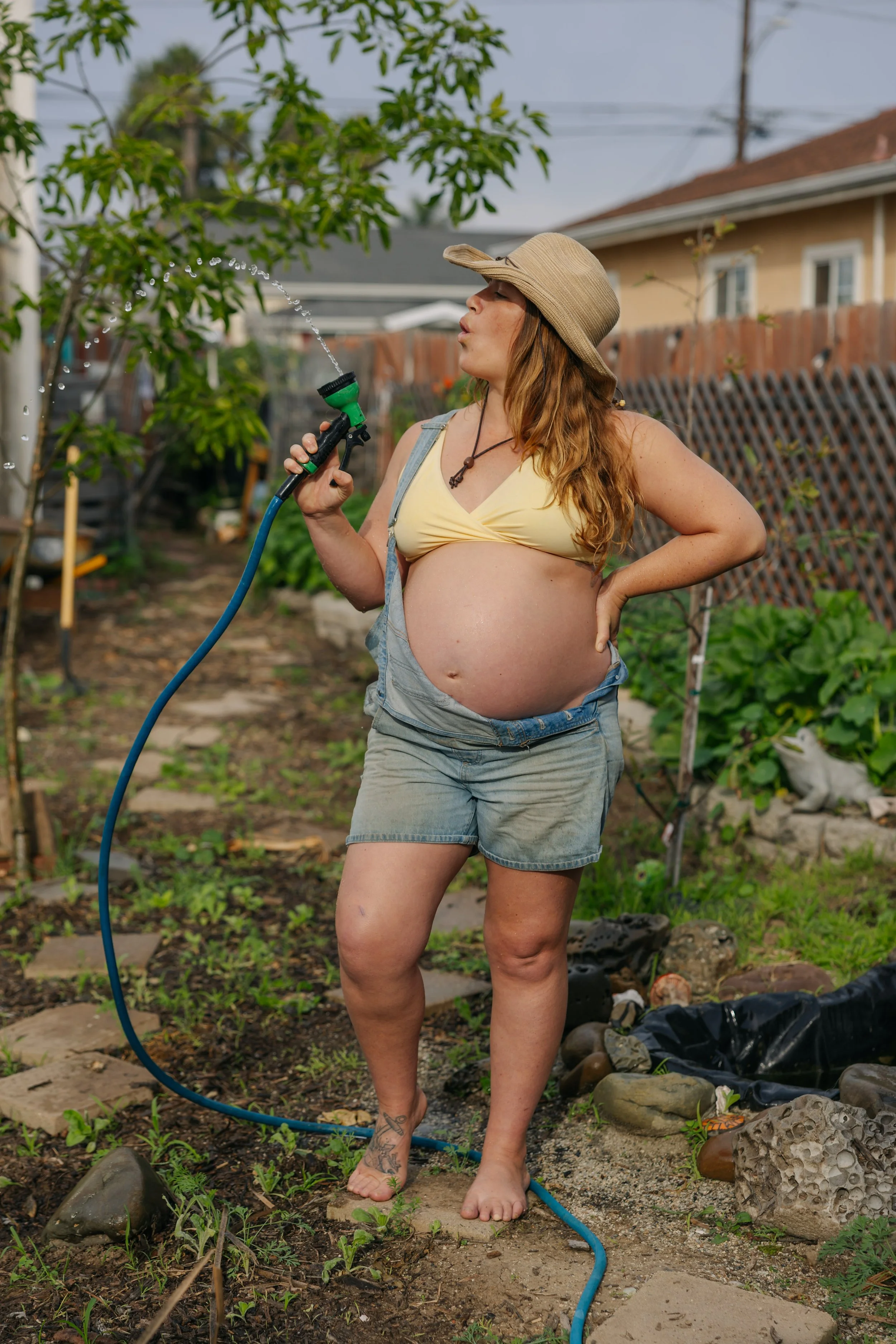A pregnant woman in a garden watering plants with a garden hose. She is wearing a beige sun hat, a yellow top, and denim shorts.