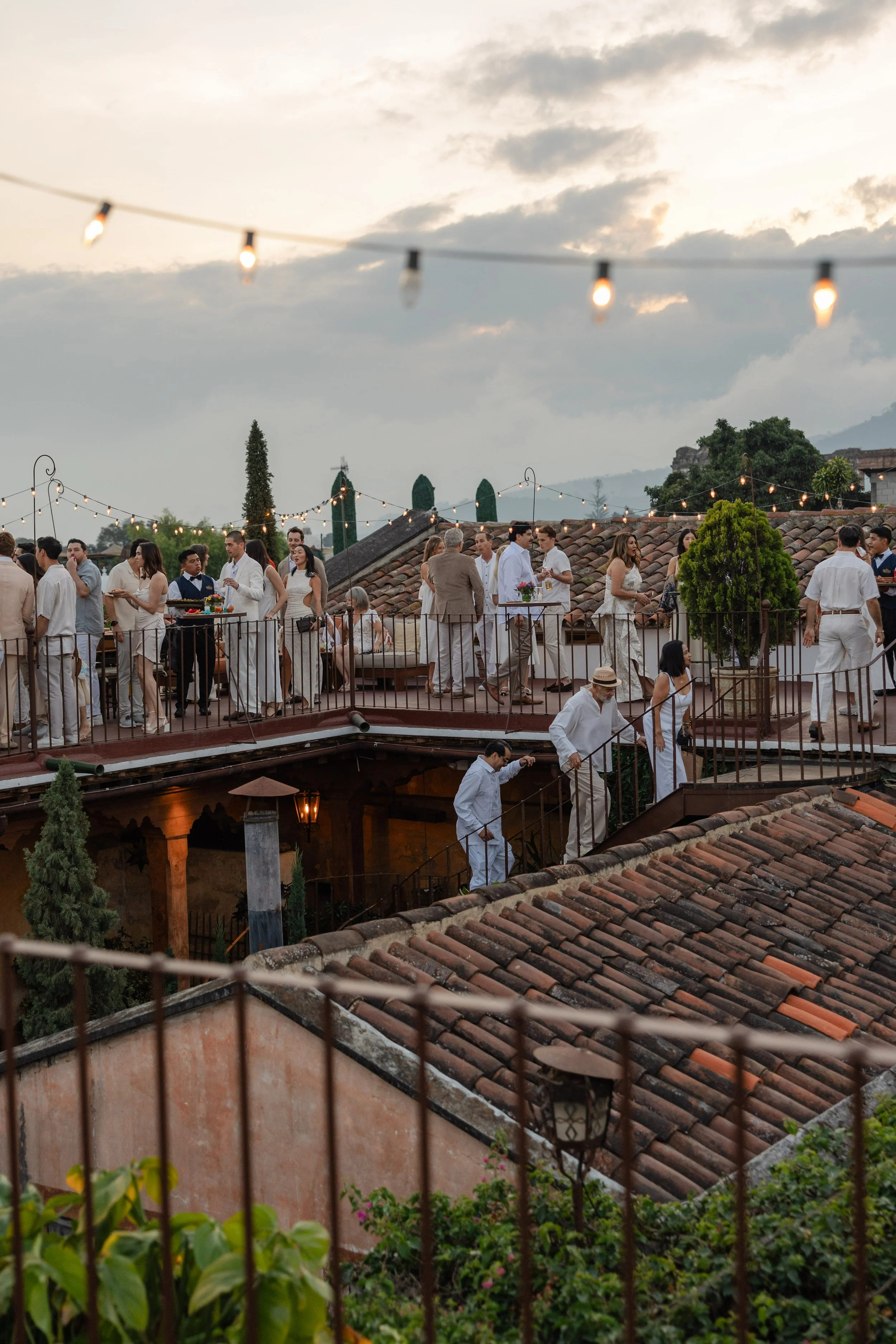 People gather on a rooftop terrace with string lights at sunset, celebrating at an outdoor event in a picturesque setting with tiled rooftops and distant mountains.