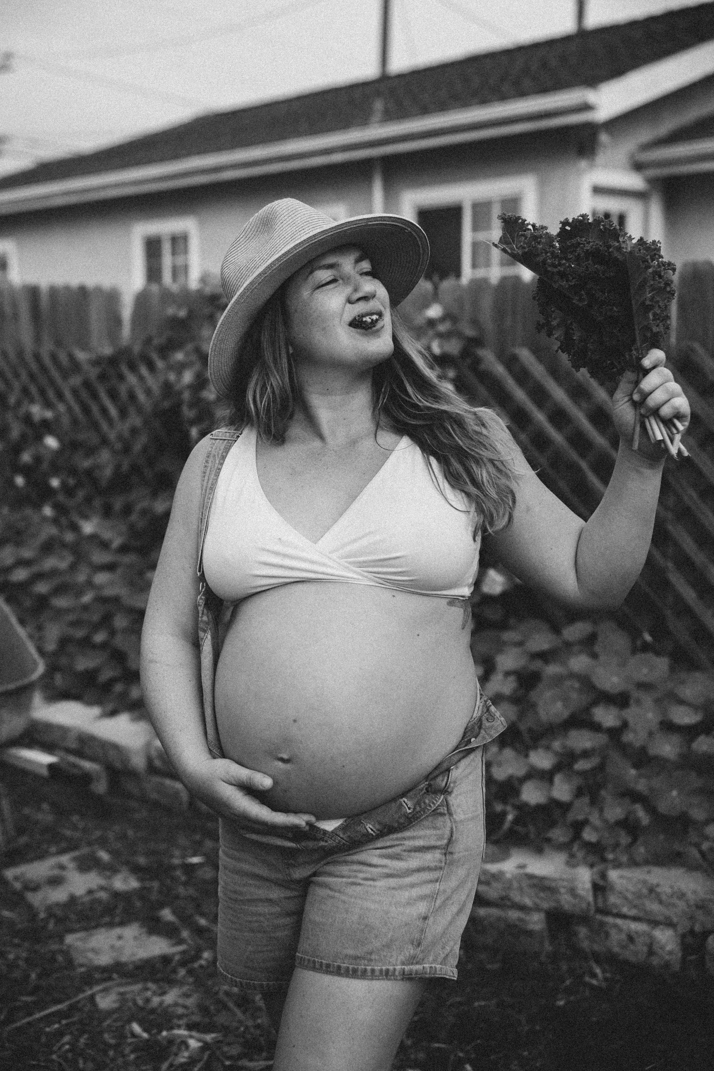 A pregnant woman wearing a hat and tank top, holding a bunch of kale outside in a garden or backyard, smiling or singing.