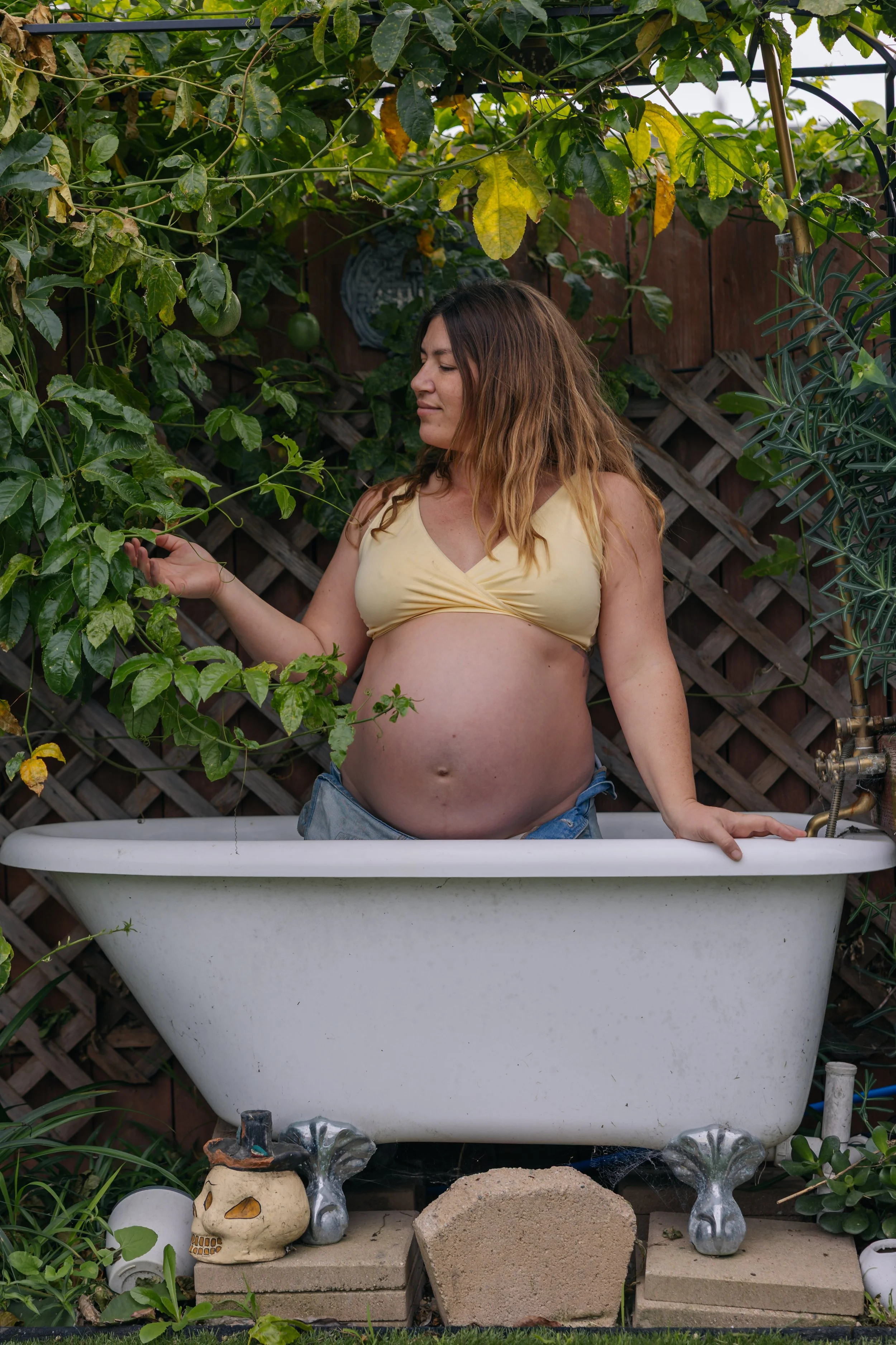 Pregnant woman standing in a white bathtub outdoors, surrounded by green plants and a wooden fence.