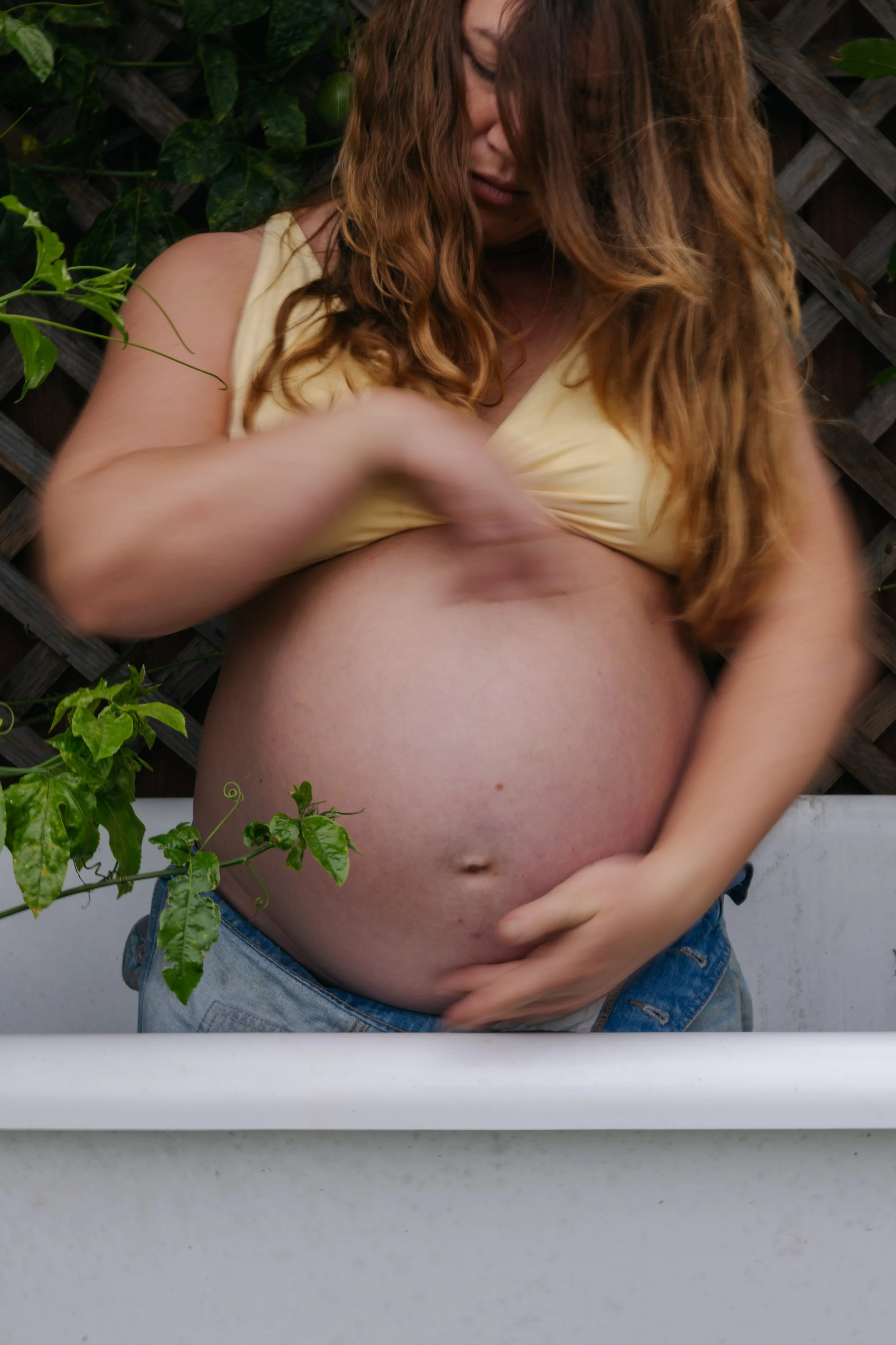 A pregnant woman with long curly hair wearing a beige top and jeans, standing outdoors in front of a wooden fence and green plants, cradling her belly with one hand.