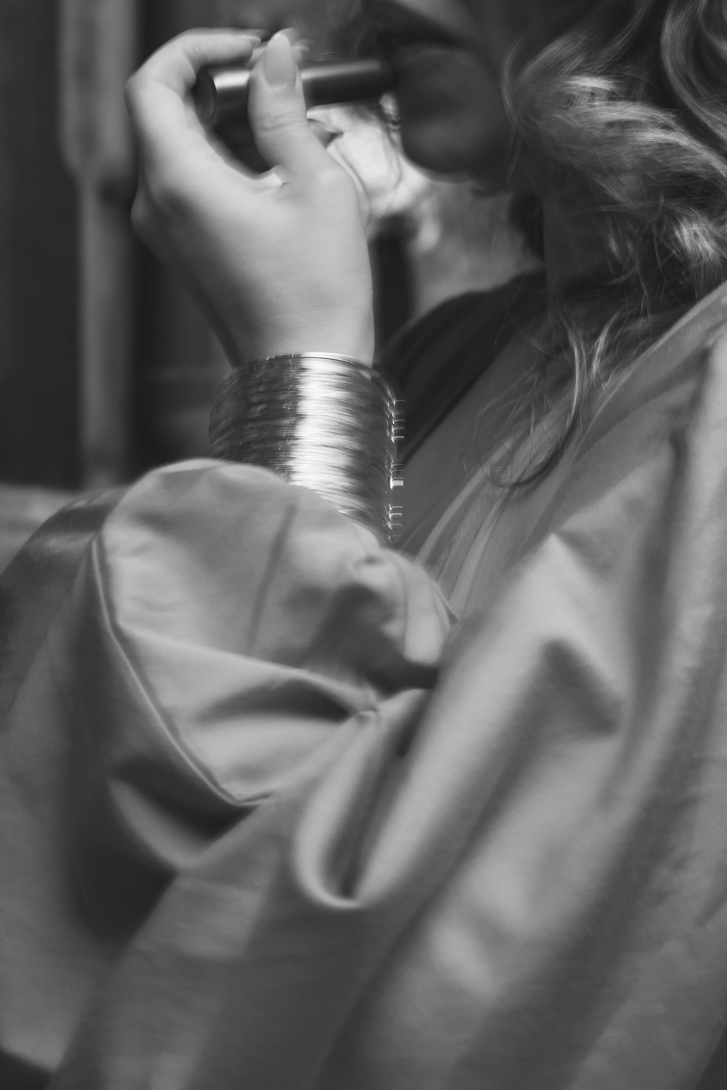 A woman with wavy hair wearing a jacket and large bangles, drinking from a glass with a straw.