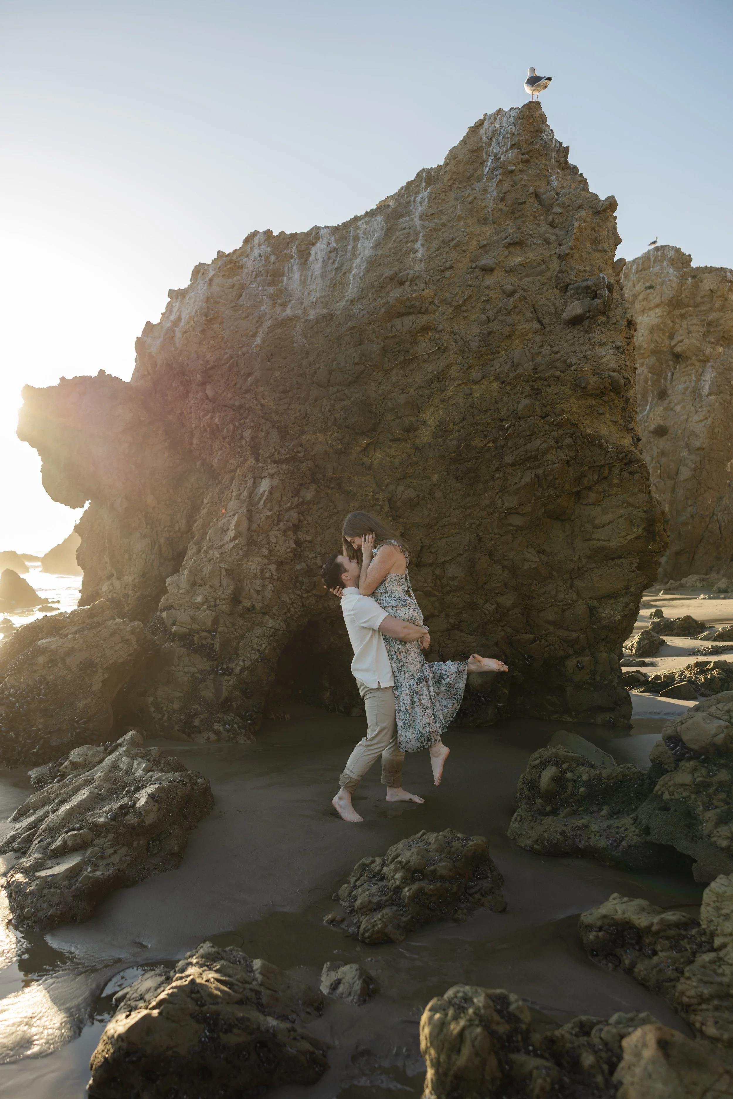 A couple embracing and kissing on the beach near large rocks and a prominent rock formation with seagulls perched on top, during sunset.