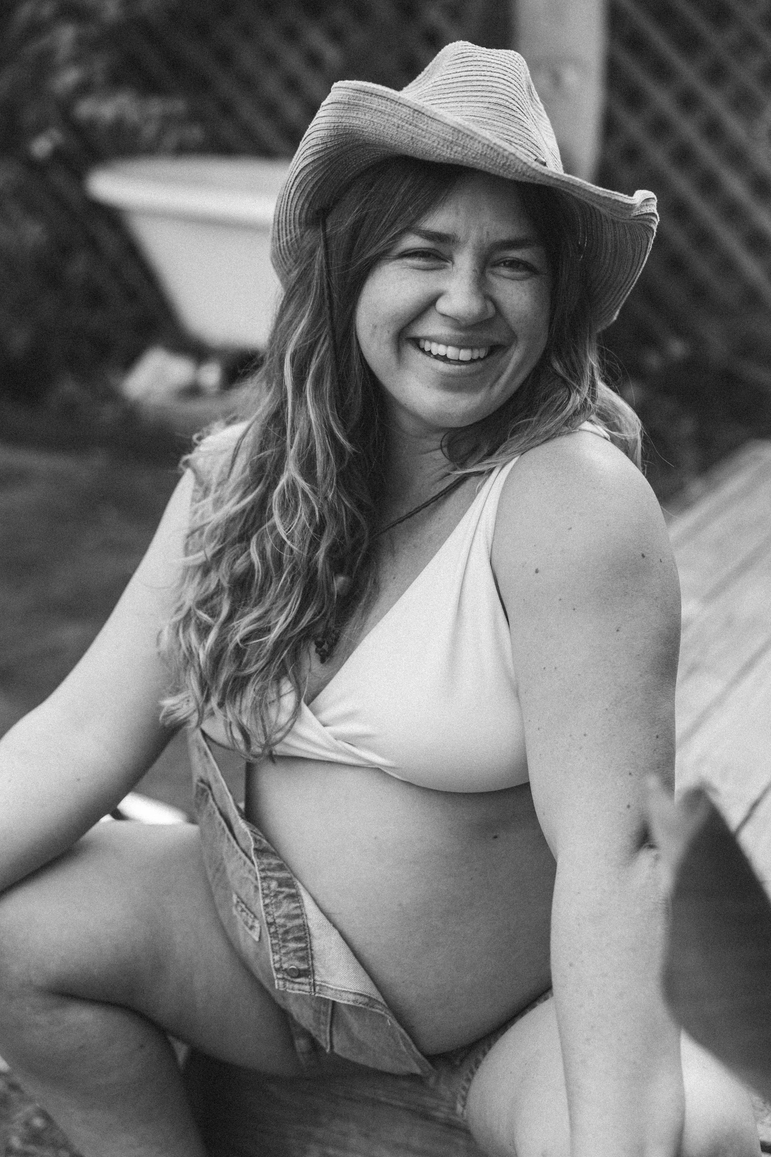 A young woman with long, curly hair wearing a wide-brimmed hat and a sleeveless top, smiling while sitting outdoors.