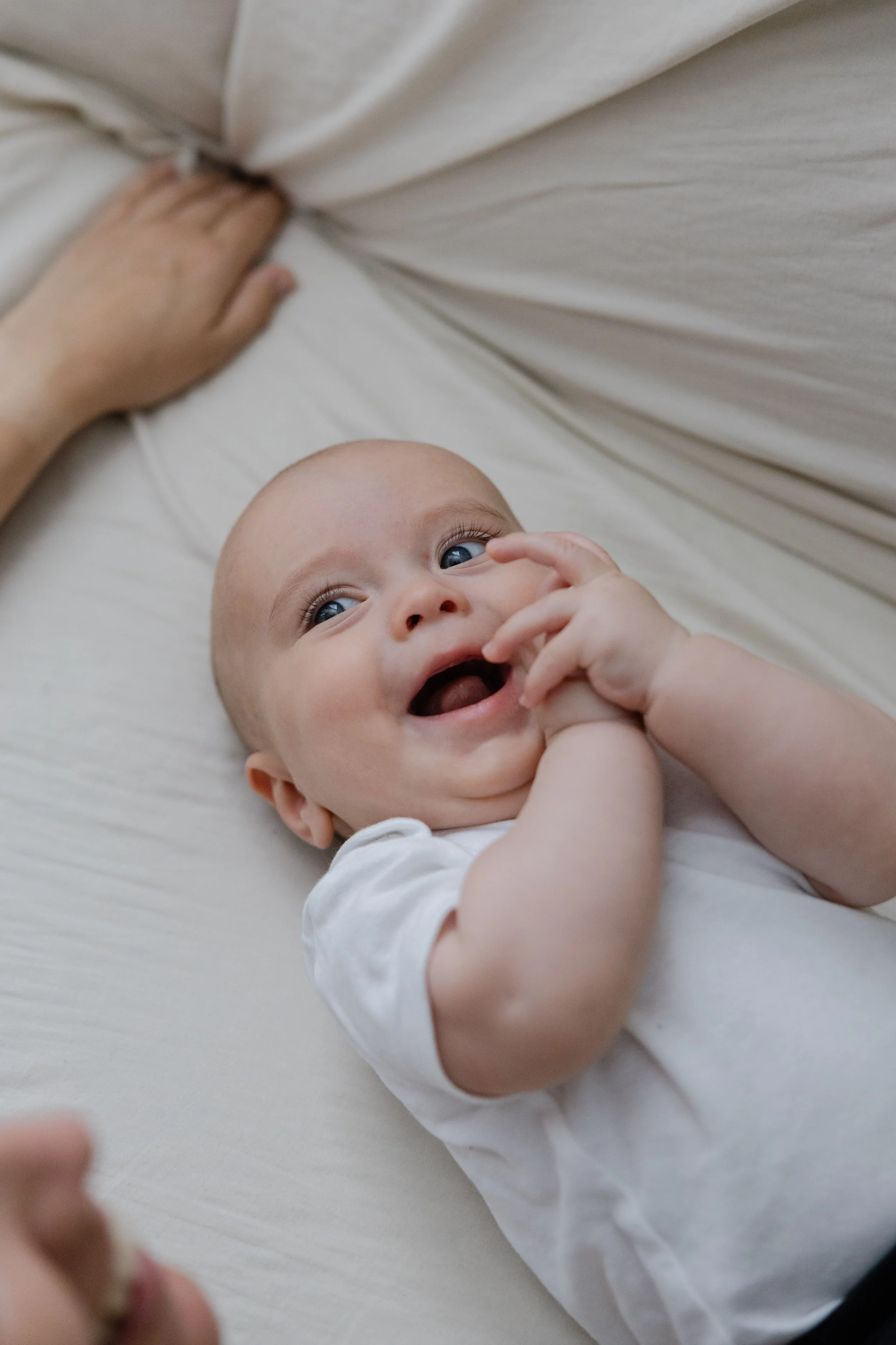 Smiling baby lying on a bed, wearing a white onesie, with a hand touching their face and a hand resting on the bed.