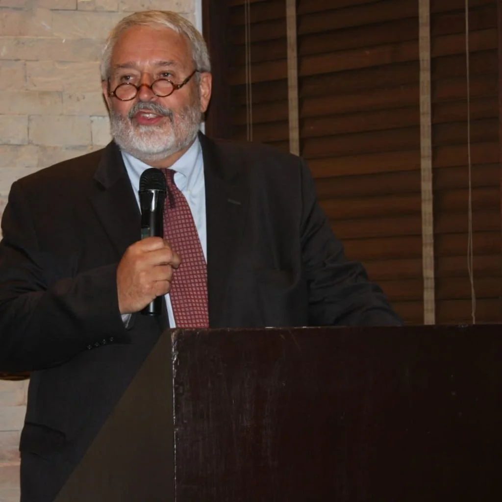 An older man with gray hair, beard, and glasses speaking into a microphone behind a dark wooden podium, wearing a black suit, light blue shirt, and red tie, in a room with a stone wall and wooden window blinds.