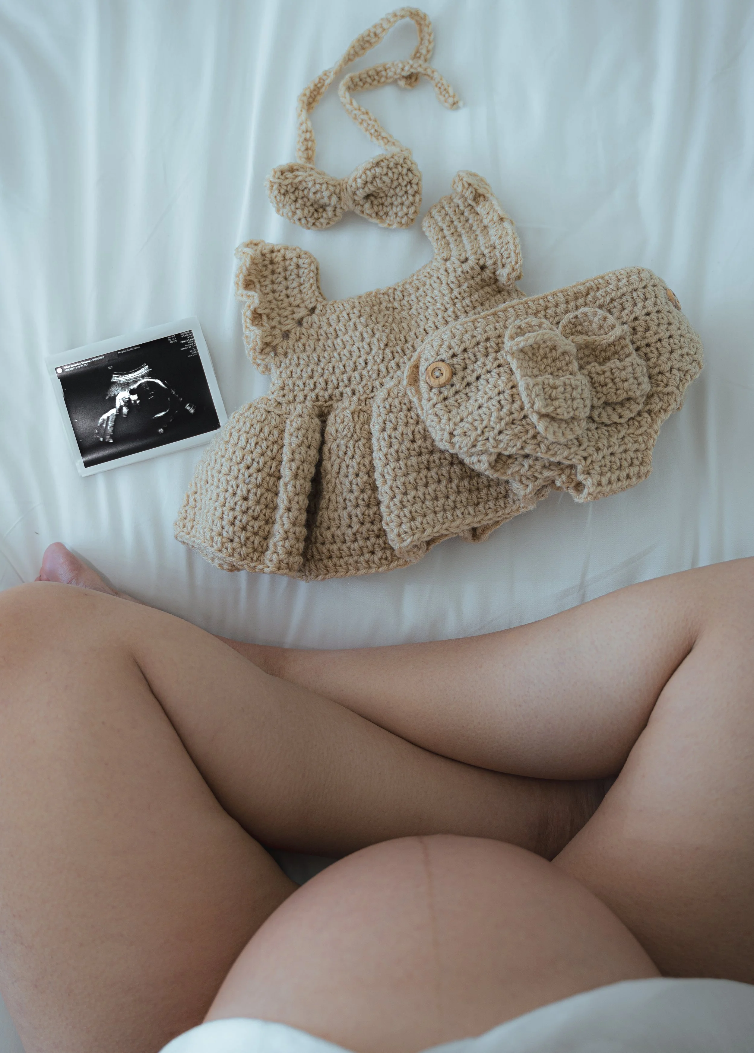 A person's legs and part of their torso are visible in the foreground, with a white bed sheet. On the bed, there are beige knitted baby clothes, including a pair of booties, a hat, and a sweater with a button. Also, there is an ultrasound image of a 