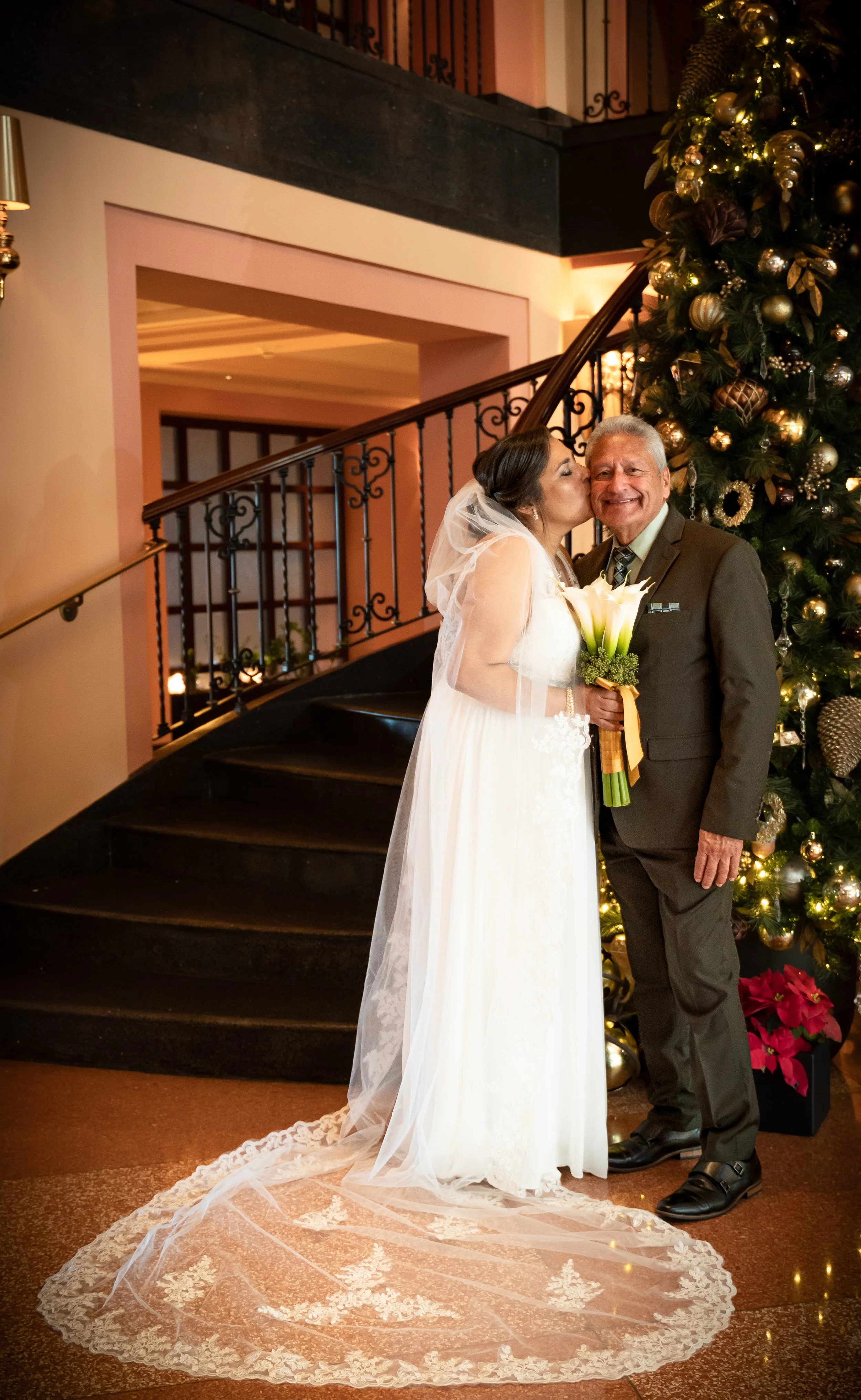 A bride in a white wedding dress and veil kisses an older man in a brown suit, holding a bouquet, in front of a decorated Christmas tree.