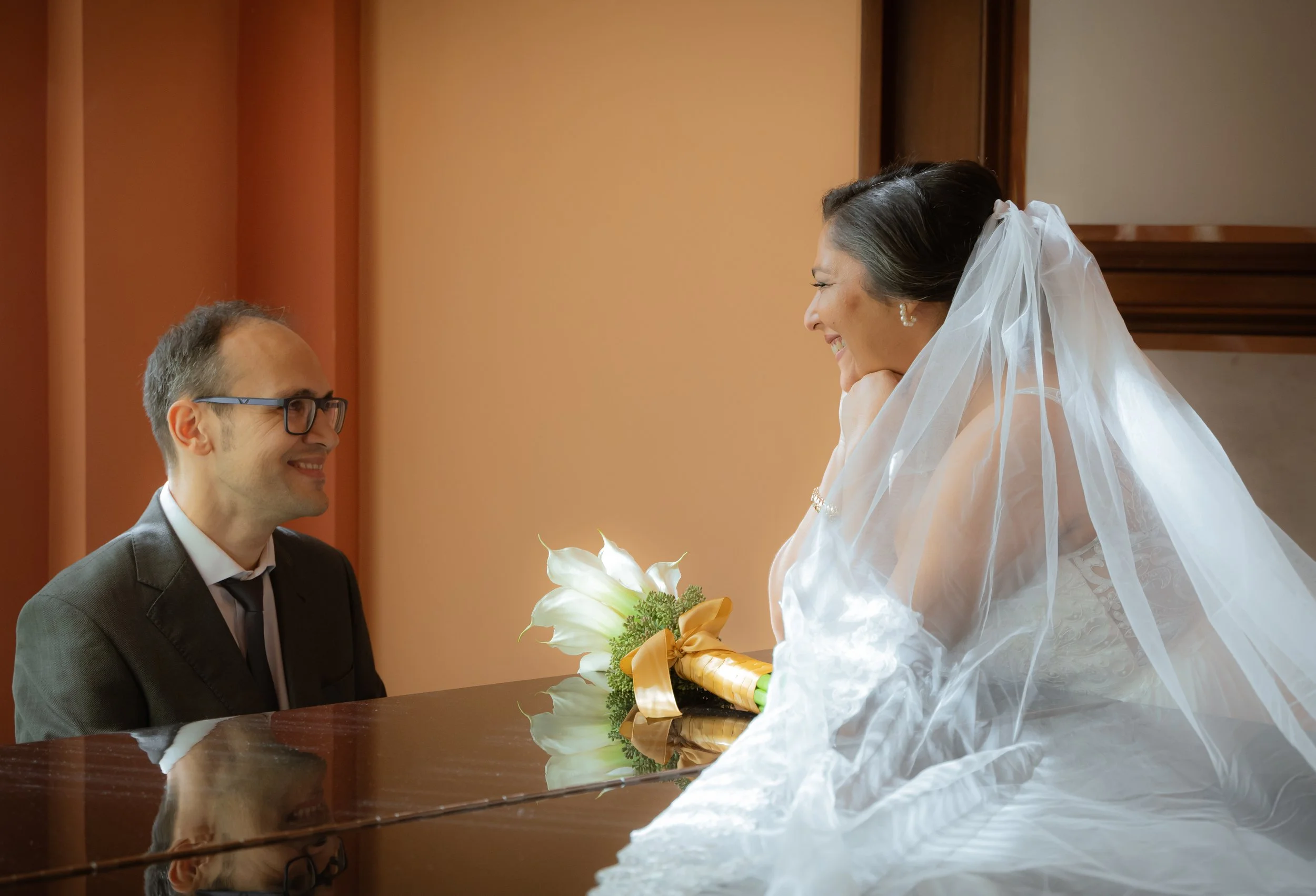 A bride and groom sharing a moment, with the bride holding a bouquet of white flowers and the groom wearing glasses, both smiling.