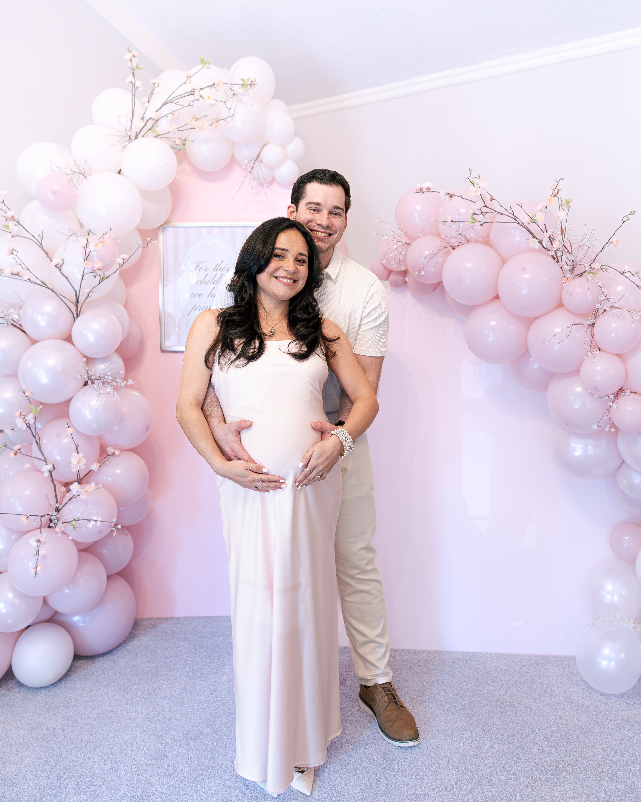 A pregnant woman in a light pink dress and a man standing behind her, both smiling, in front of a pink balloon and branch decoration backdrop.