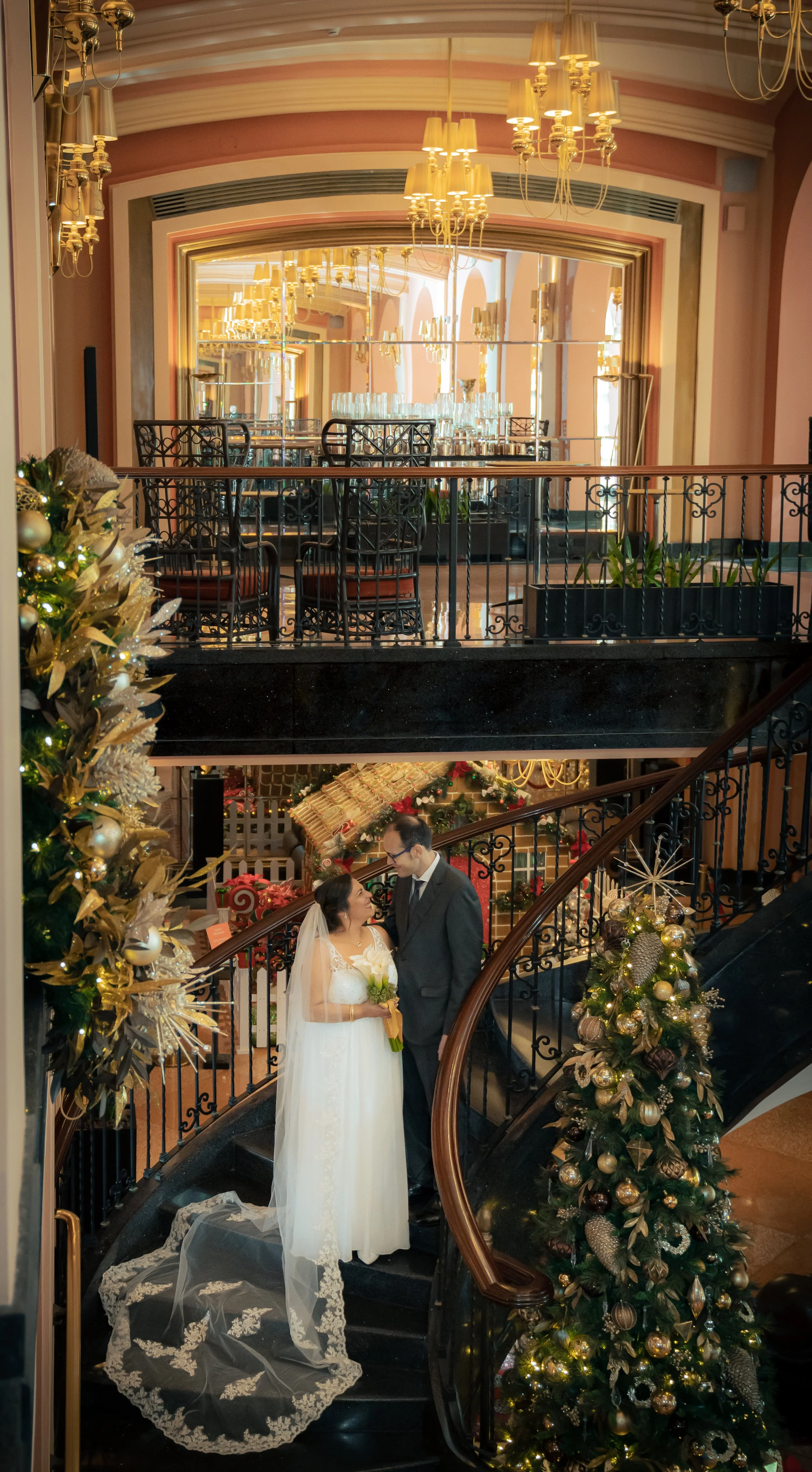 A bride and groom stand on a staircase decorated with Christmas ornaments, looking at each other inside a festively decorated venue with Christmas trees and chandeliers.