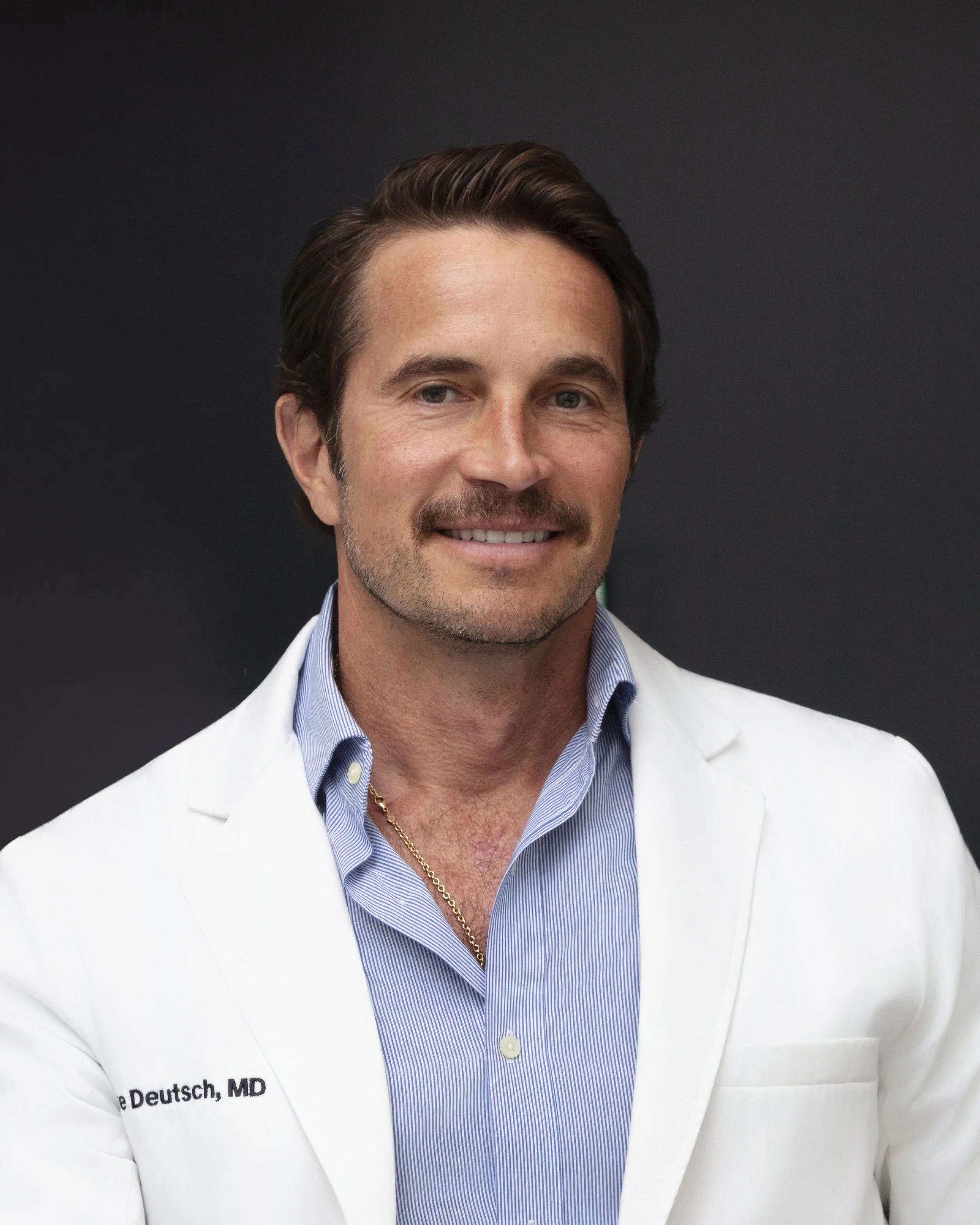 A man in a white lab coat with a blue shirt underneath, smiling, against a dark background.
