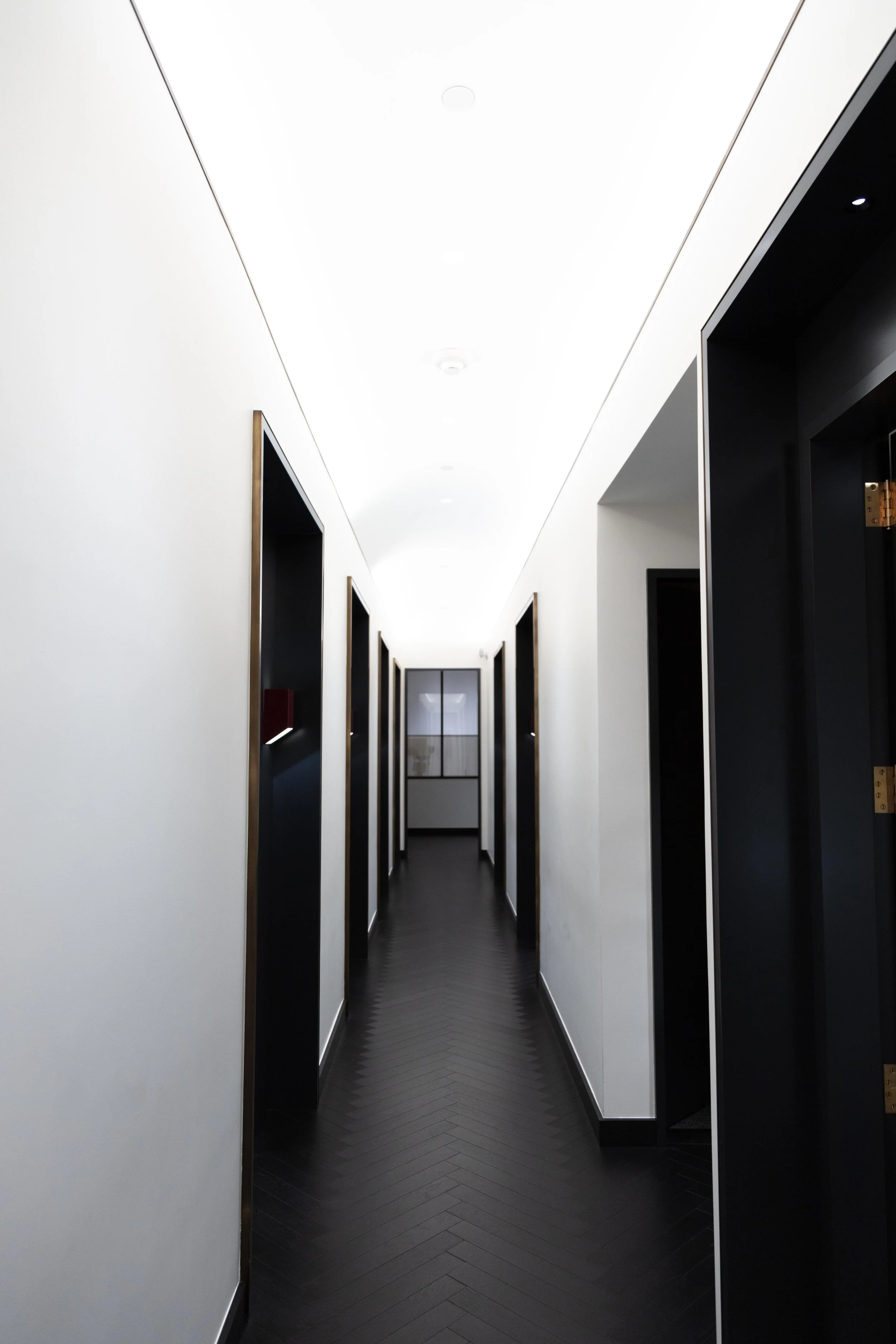 A modern, minimalist hallway with white walls, black doors, and dark wooden herringbone flooring. Light fixtures are recessed in the ceiling, and there is a window at the end of the hallway.