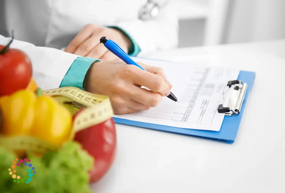 A healthcare professional holds a blue pen and is filling out a form on a blue clipboard. In the foreground, there are apples, a yellow bell pepper, and a red bell pepper with a measuring tape wrapped around them.