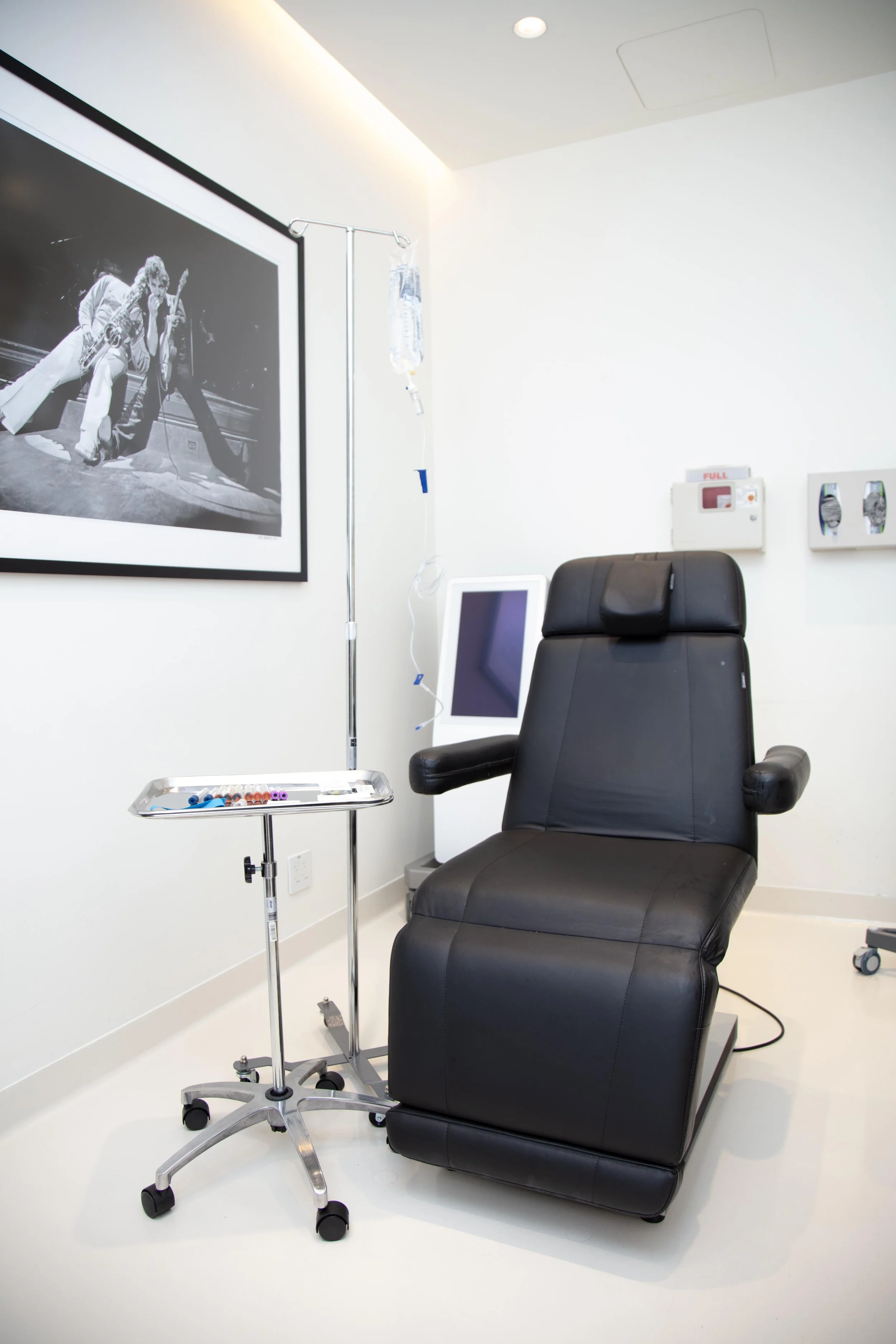 Hospital room with black reclining chair, IV stand, tray with syringes, and medical monitor, framed black and white photograph of rock performers on wall.