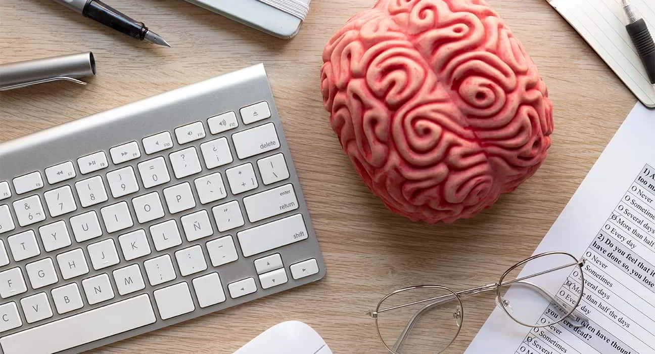 A desk with a computer keyboard, a spherical object resembling a brain, eyeglasses, pens, a notebook, and papers on a wooden surface.