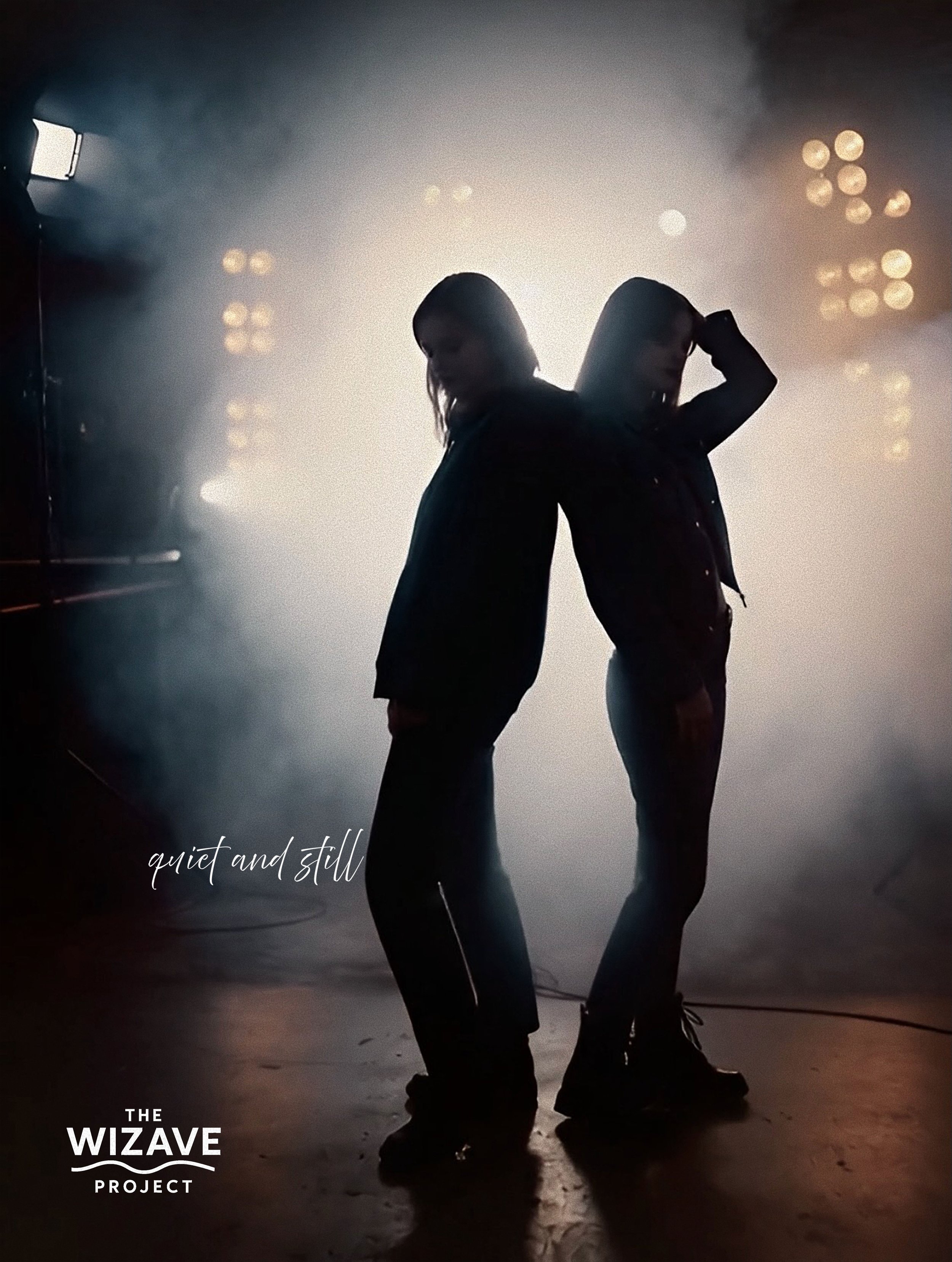 Silhouettes of two people back-to-back in a dimly lit setting with fog and stage lights, with text saying 'quiet and still' and 'The Wizave Project'.