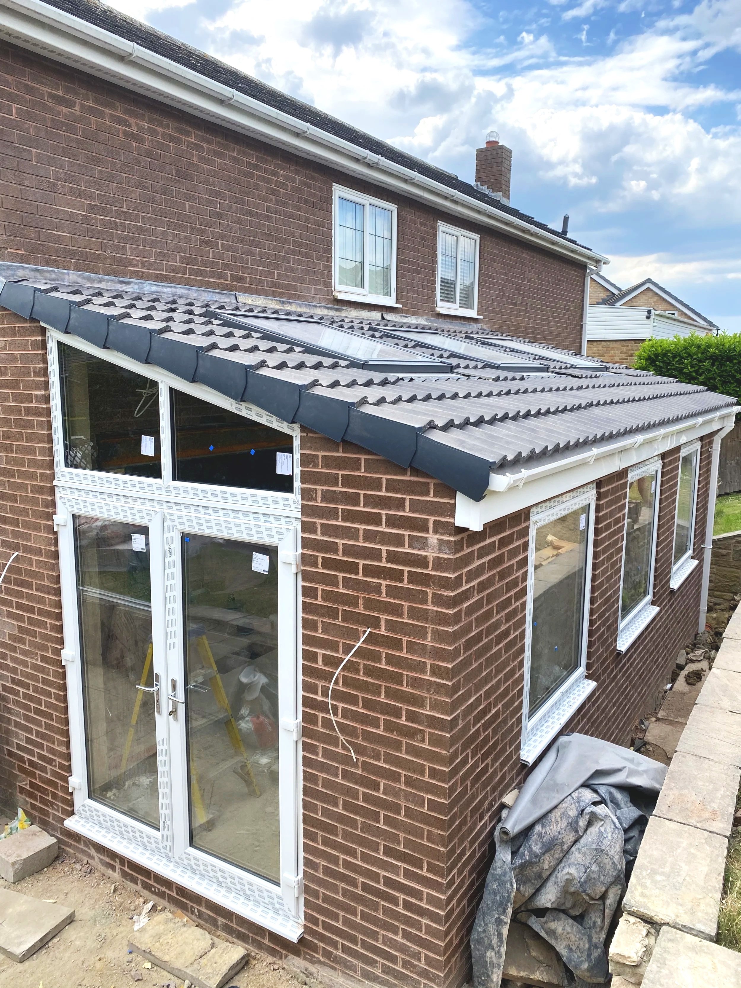 A brick house with a conservatory under construction, featuring a tiled roof, white window frames, and a glass door, with construction materials and a cloudy sky in the background.