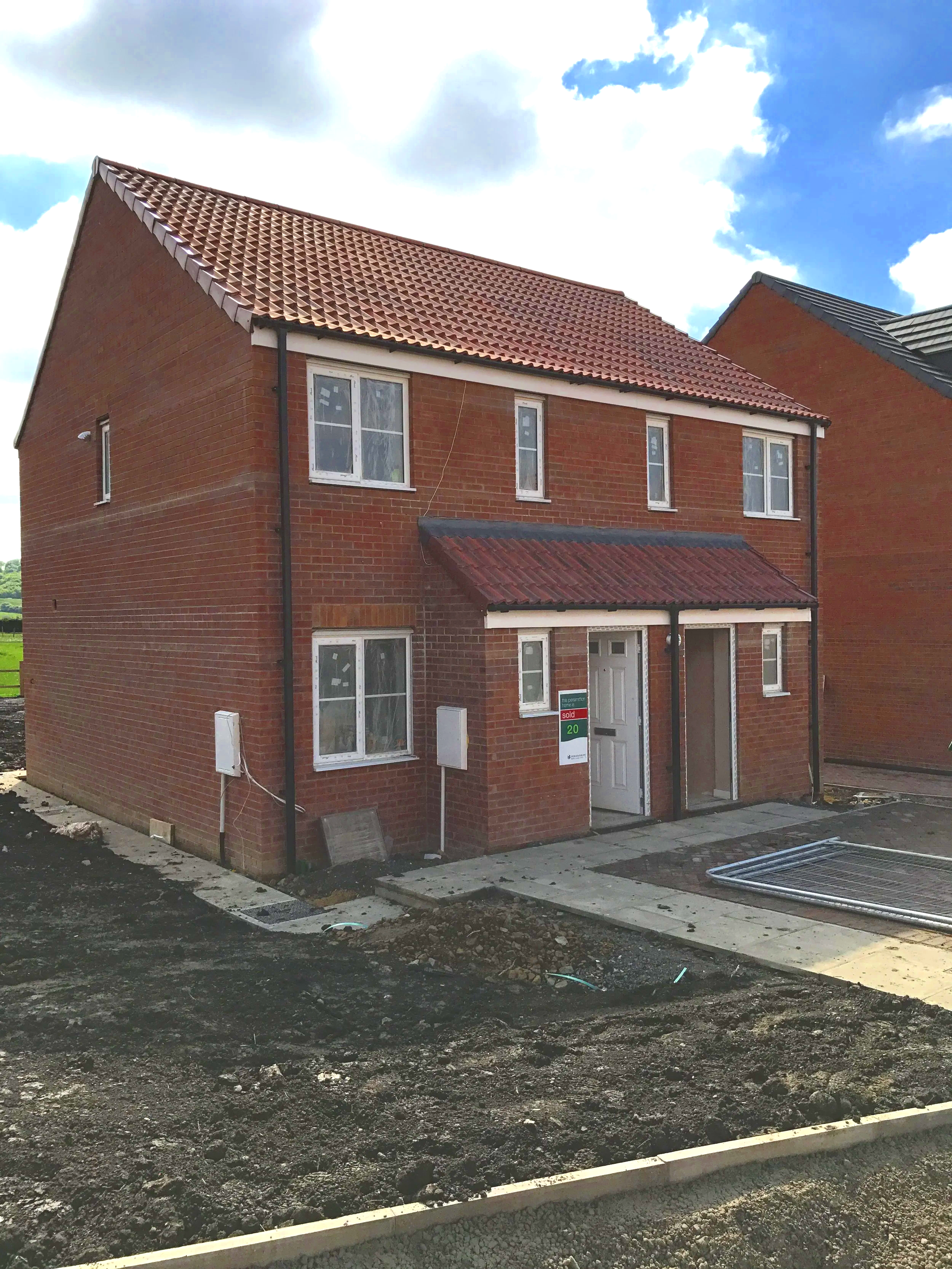 Newly built brick house with a red tiled roof and white window frames, situated in a developing neighborhood.