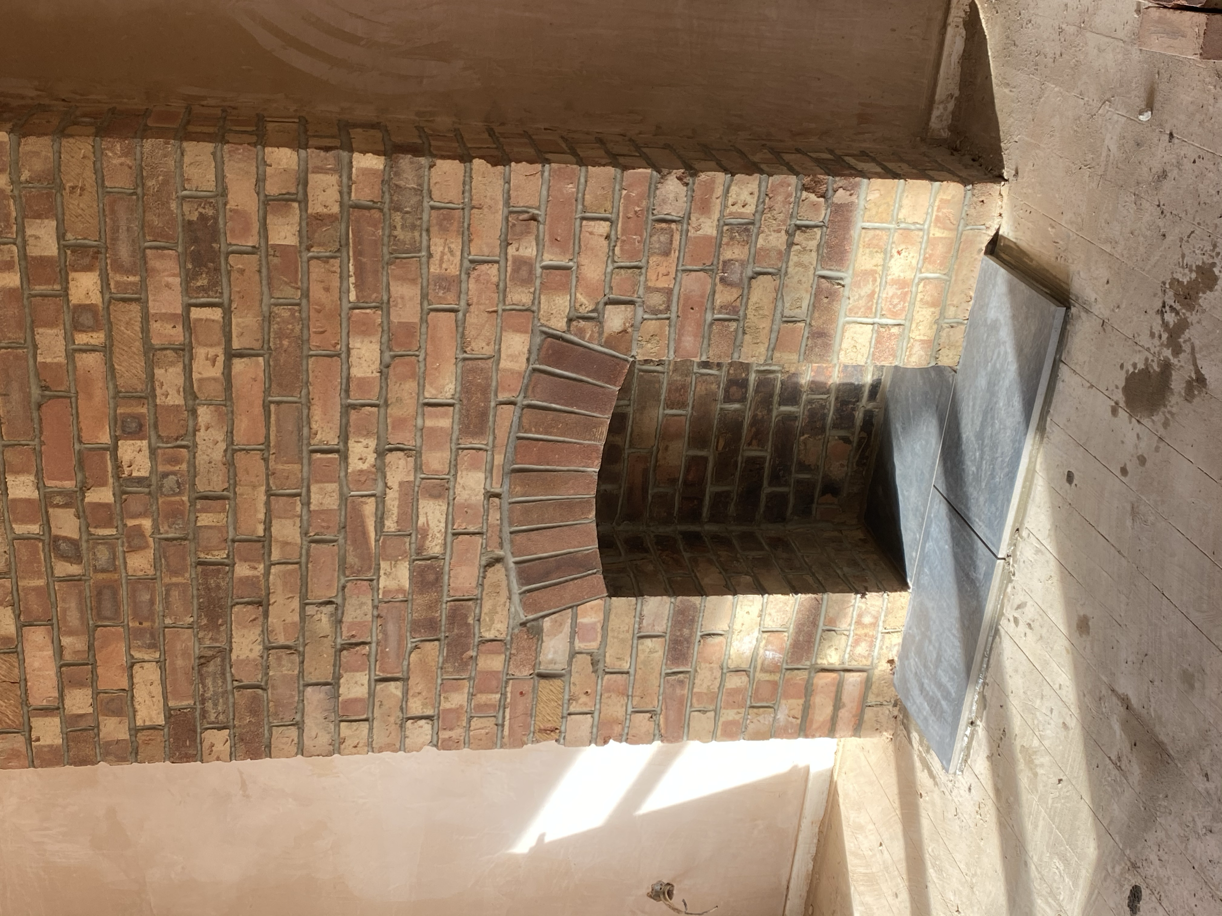 Brick fireplace with a small arched opening and metal fire grate inside, under construction in a room with wooden floor.