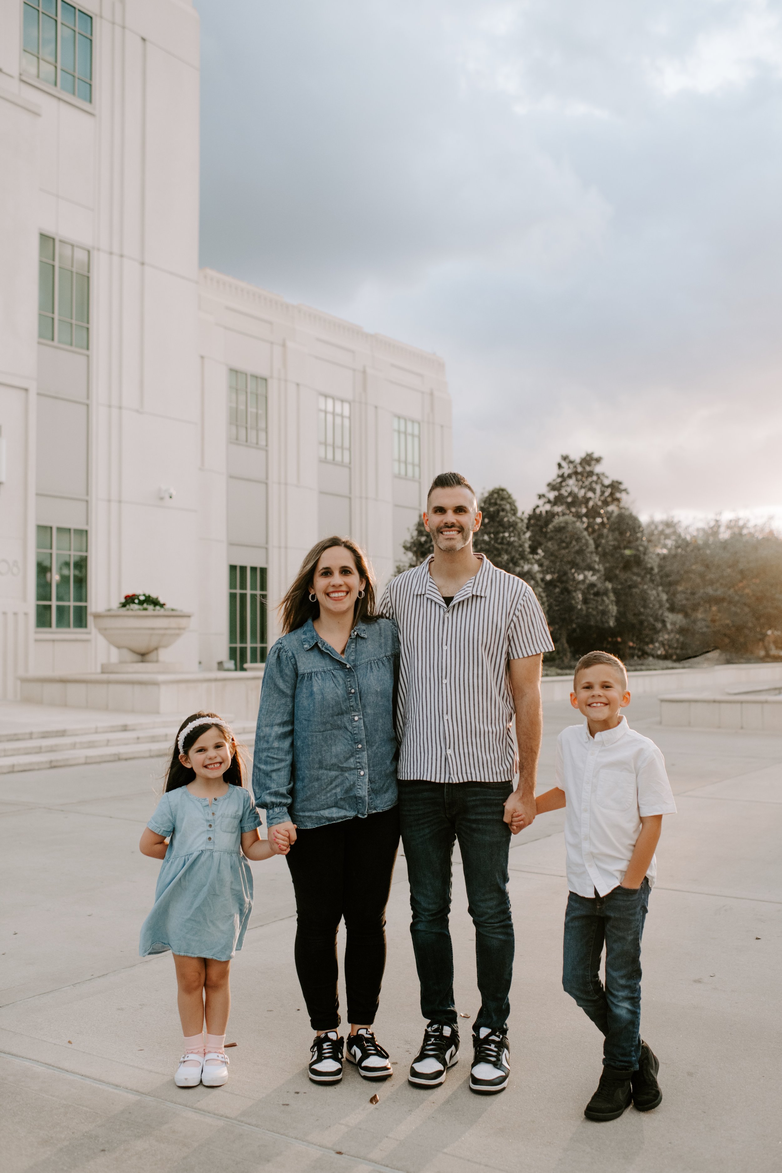 A family of four holding hands outside a white building with steps, with trees and a cloudy sky in the background.