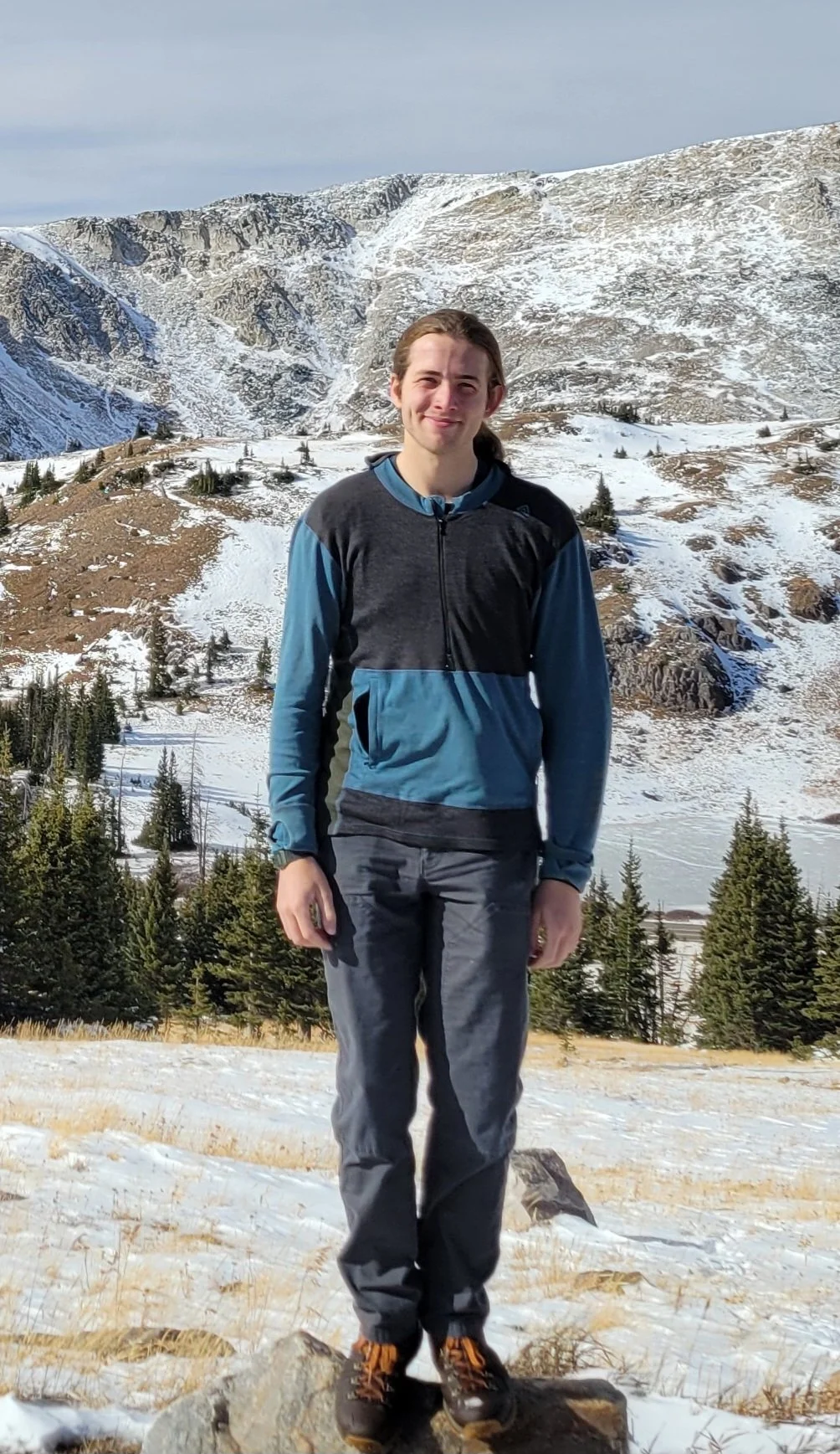 A young man standing on a rock in a snowy mountain landscape with trees and mountains in the background.
