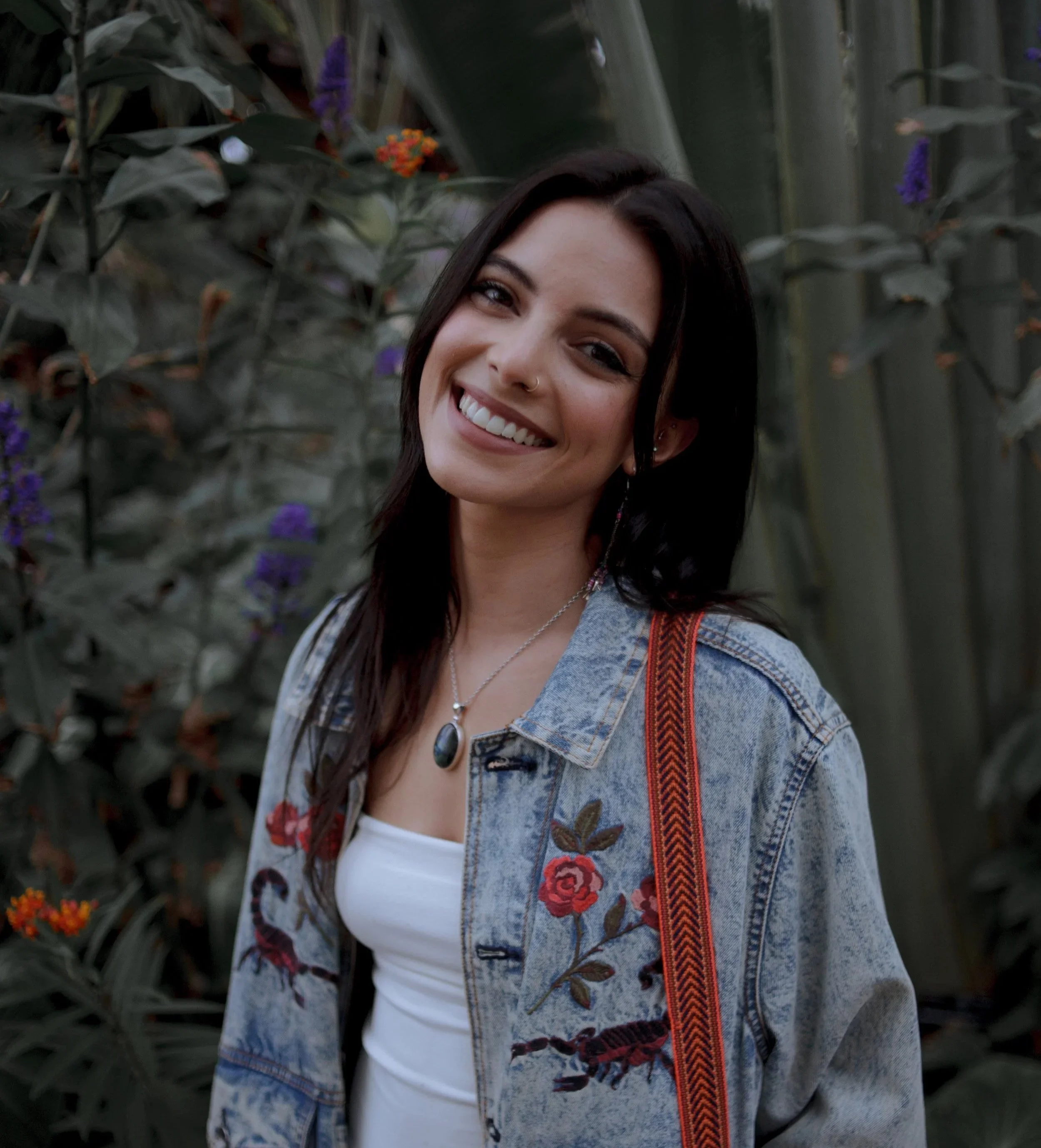 A young woman with dark hair smiling outdoors, wearing a denim jacket with embroidered flowers and a white top, carrying a colorful strap bag, standing in front of green plants and flowers.