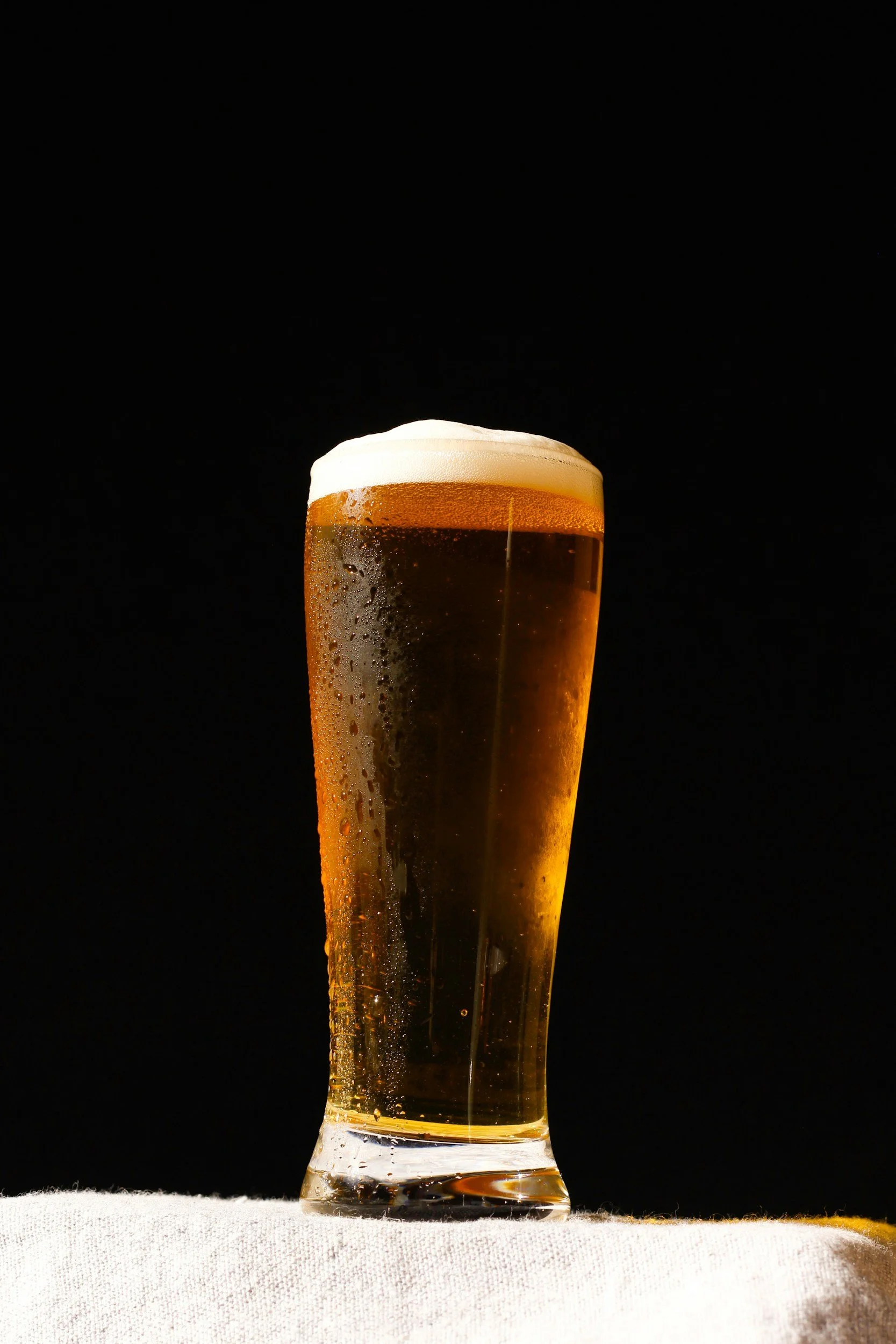 A tall glass of beer with foam on top on a white surface against a black background.