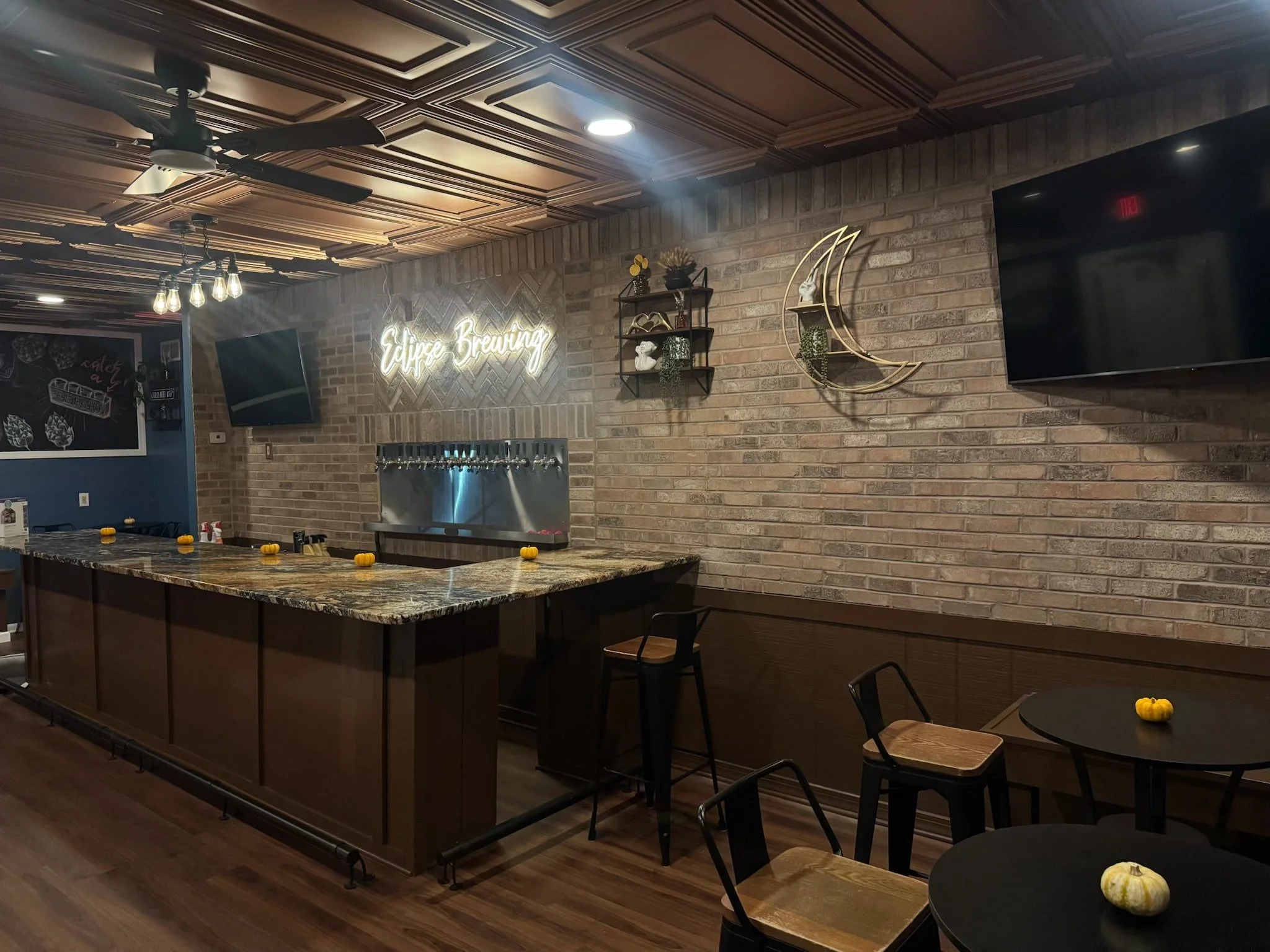 Interior of a brewery bar with exposed brick walls, a neon sign reading 'Eclipse Brewing,' a black chalkboard menu, small decorative pumpkins on tables, and a large flat-screen TV.