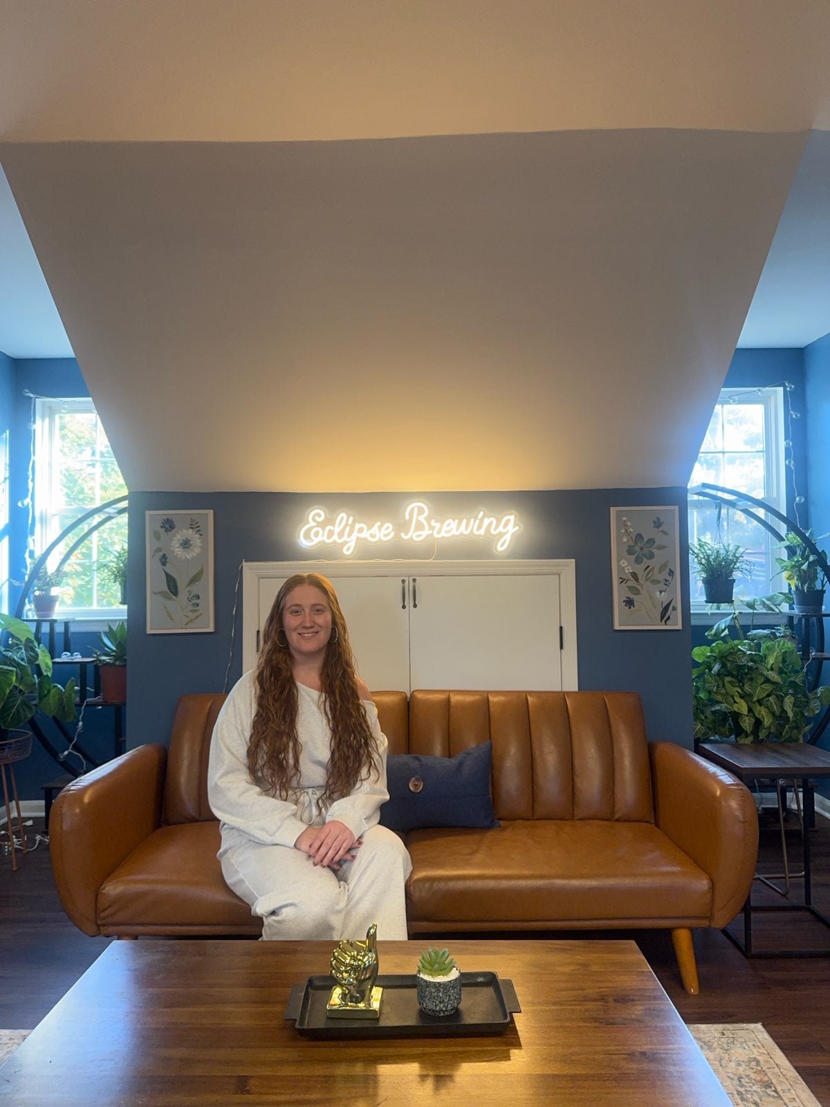 Eclipse Brewing owner Meg Hilbert, a woman, with long, wavy hair sitting on a leather couch in a cozy room with plants and windows. There is a neon sign reading 'Eclipse Brewing' on the wall behind her.