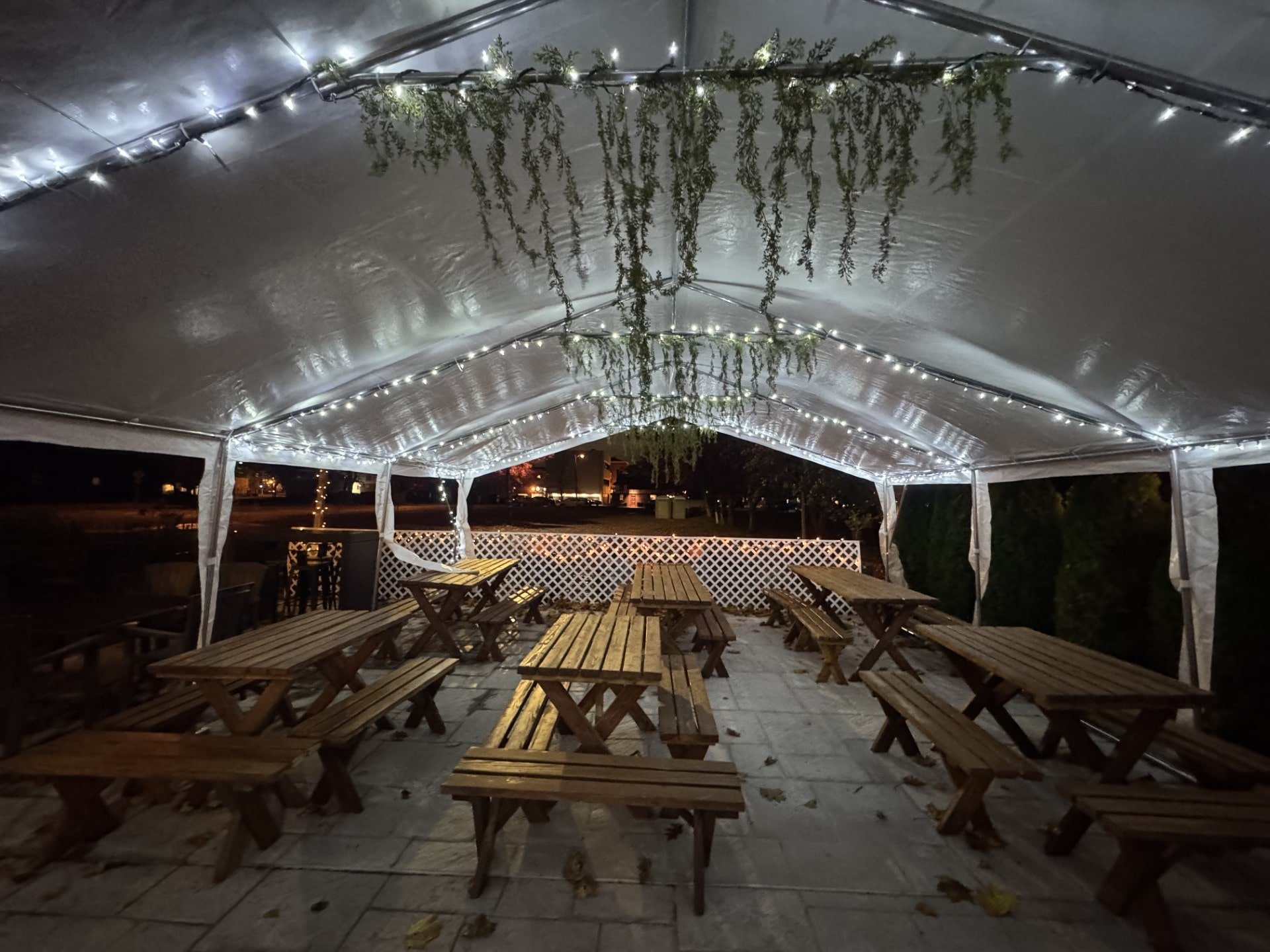 Outdoor patio area under a white canopy tent with string lights and hanging greenery, wooden picnic tables and benches, nighttime setting, available for private events at Eclipse Brewing