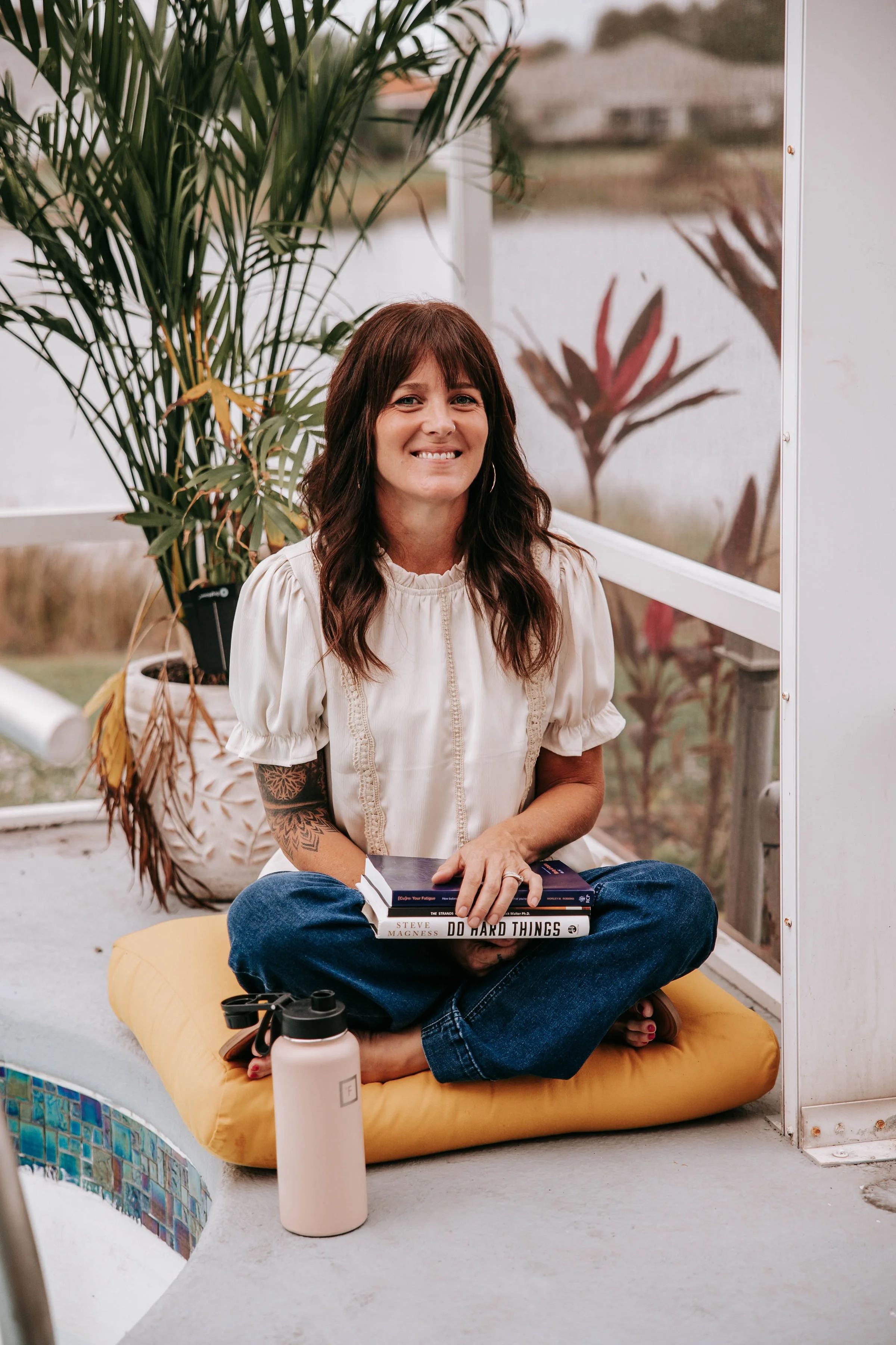 Woman sitting cross-legged on a yellow cushion, holding a stack of books, with a large potted plant behind her, in a sunroom or enclosed porch with large windows and a view of the outdoors.