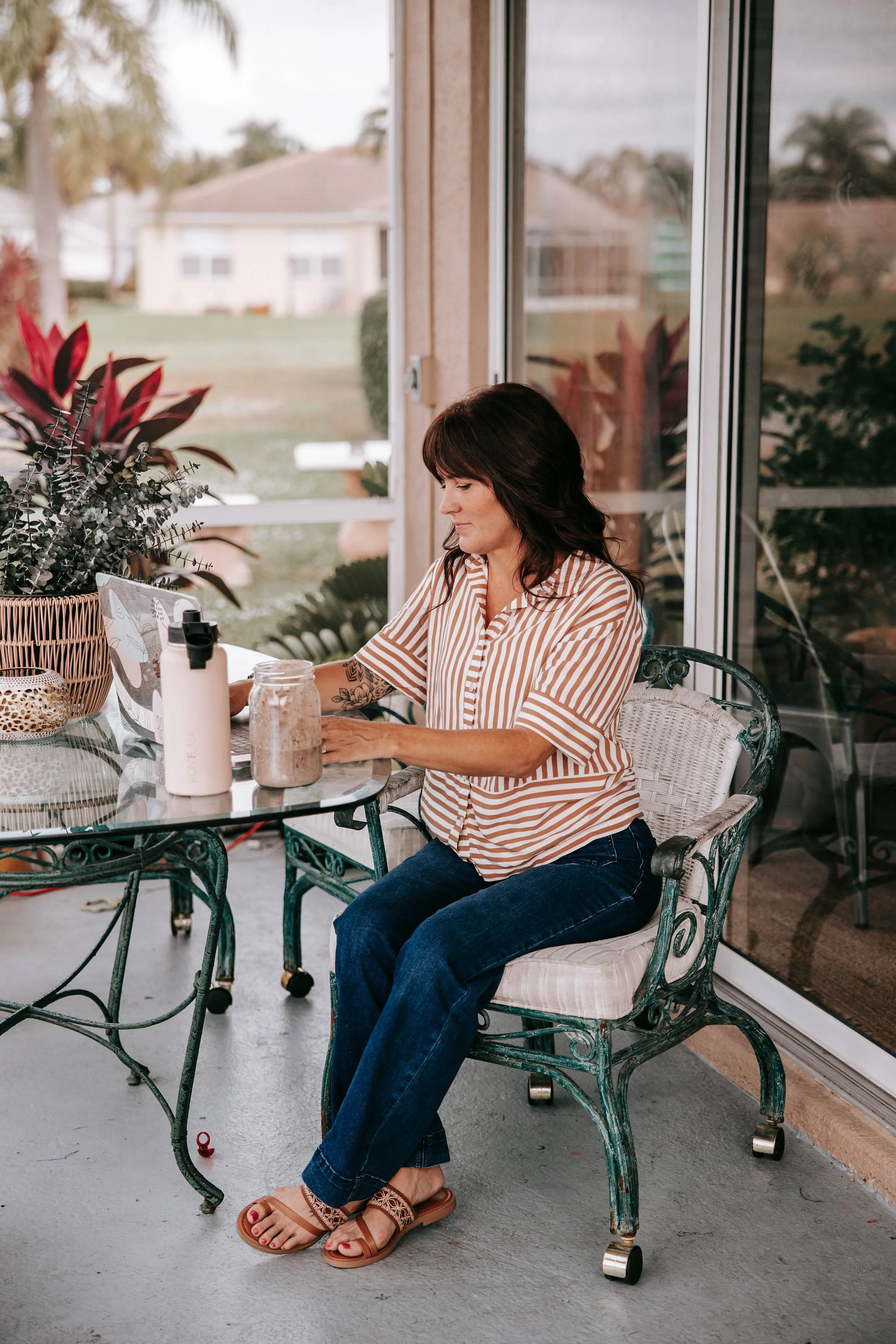 Woman sitting at an outdoor table on a porch, working on a laptop. The table has plants, a water bottle, and a jar with brown substance. The woman wears a striped shirt, jeans, and sandals.