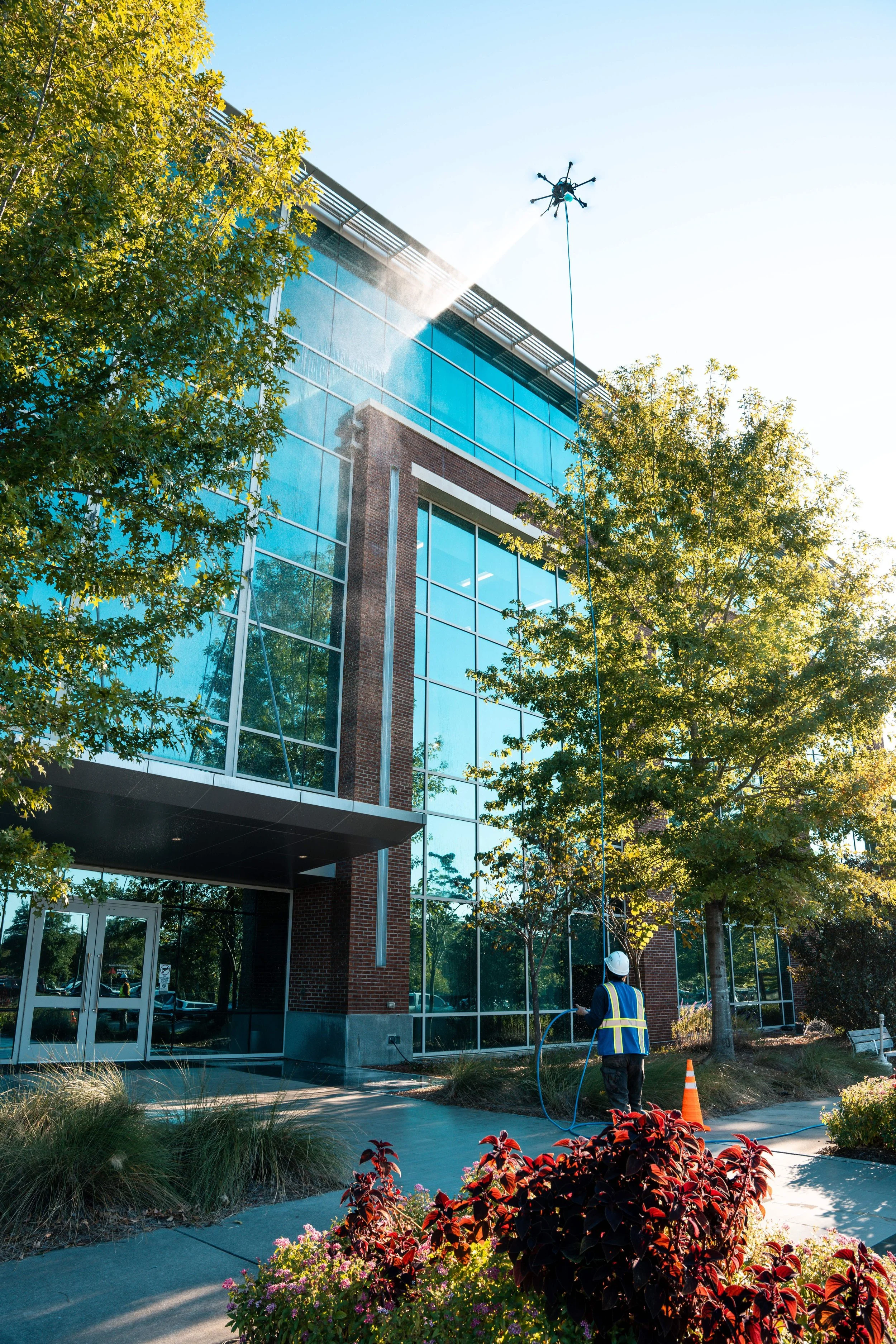 Worker in safety gear pressure washing the exterior glass windows of a modern office building with trees and plants nearby during daytime.