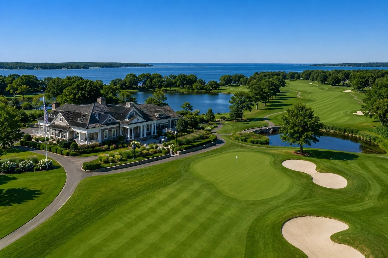 A scenic golf course with green fairways, sand bunkers, and a clubhouse surrounded by trees, with a lake and water in the background under a clear blue sky.