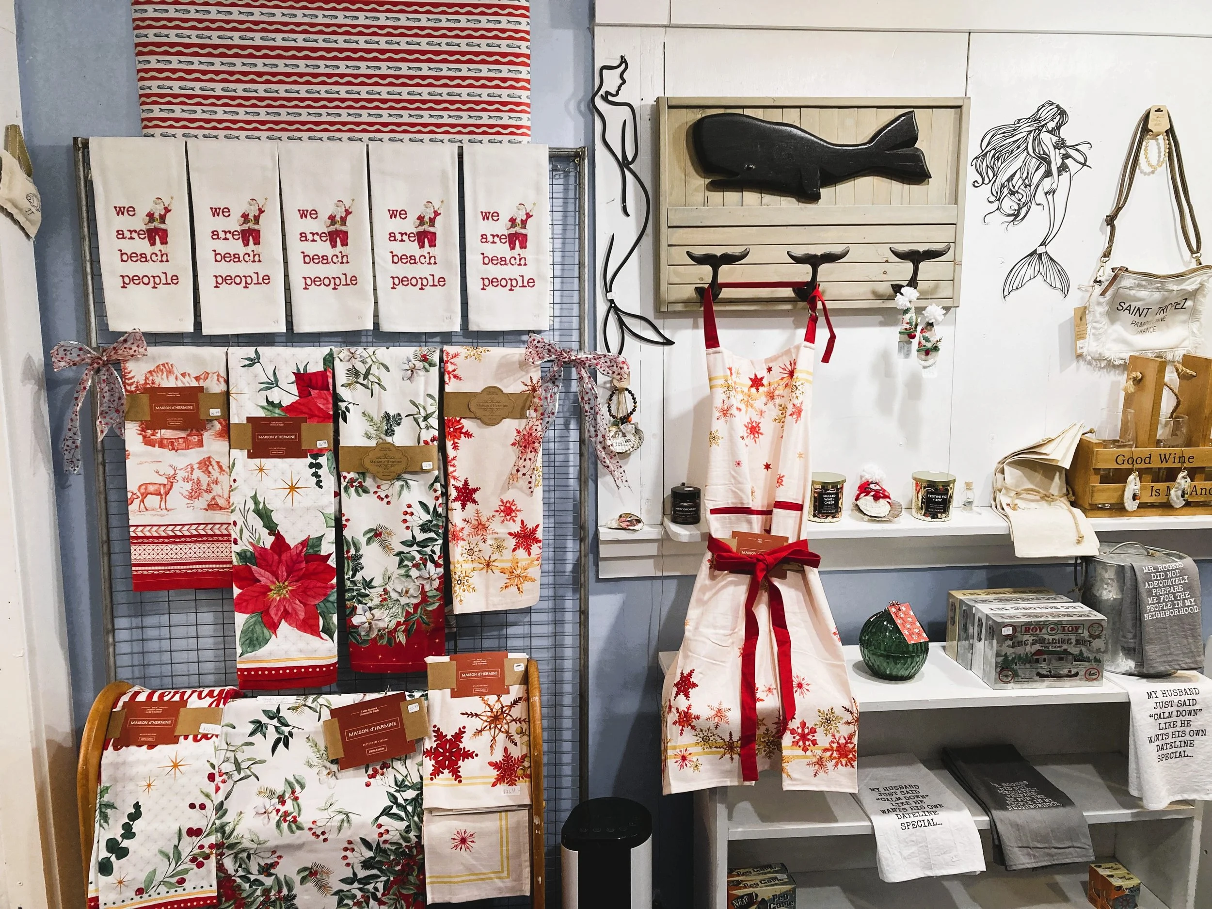 A holiday-themed display with Christmas towels, aprons, and wall decorations including a mermaid illustration and a whale sculpture. Some shelves hold candles, jars, and small decorative items.