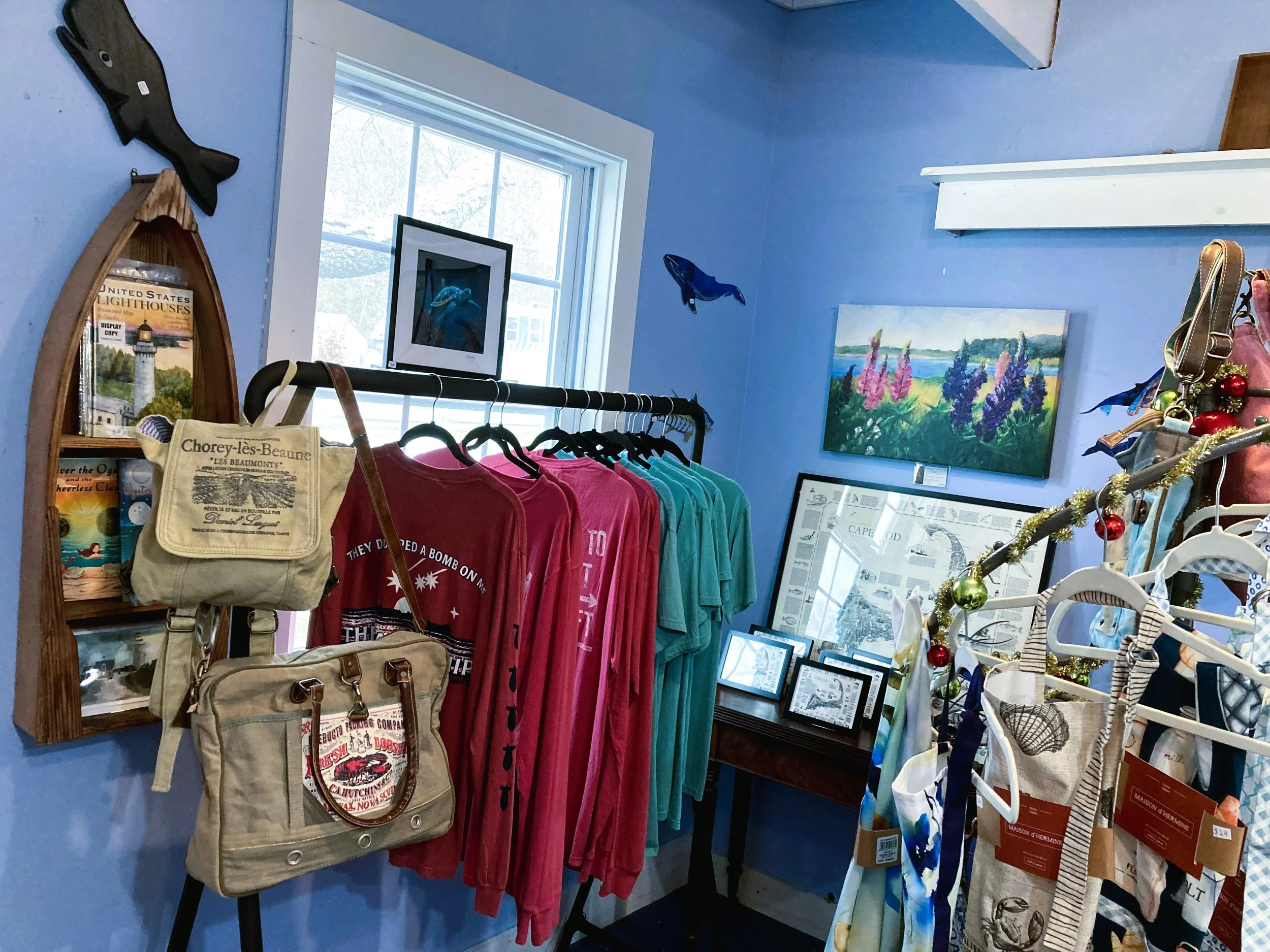 A small boutique store with blue walls displaying framed pictures, painted artwork, and iron rack with colorful T-shirts and hanging tote bags adorned with holiday ornaments.